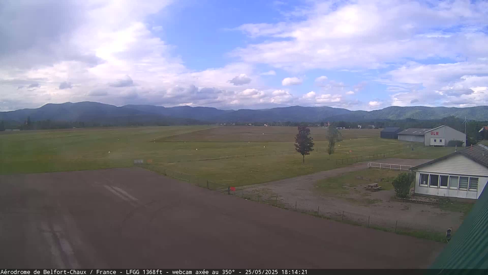 A small airfield with a runway and buildings is situated in a grassy field, with mountains in the background under a partly cloudy sky.