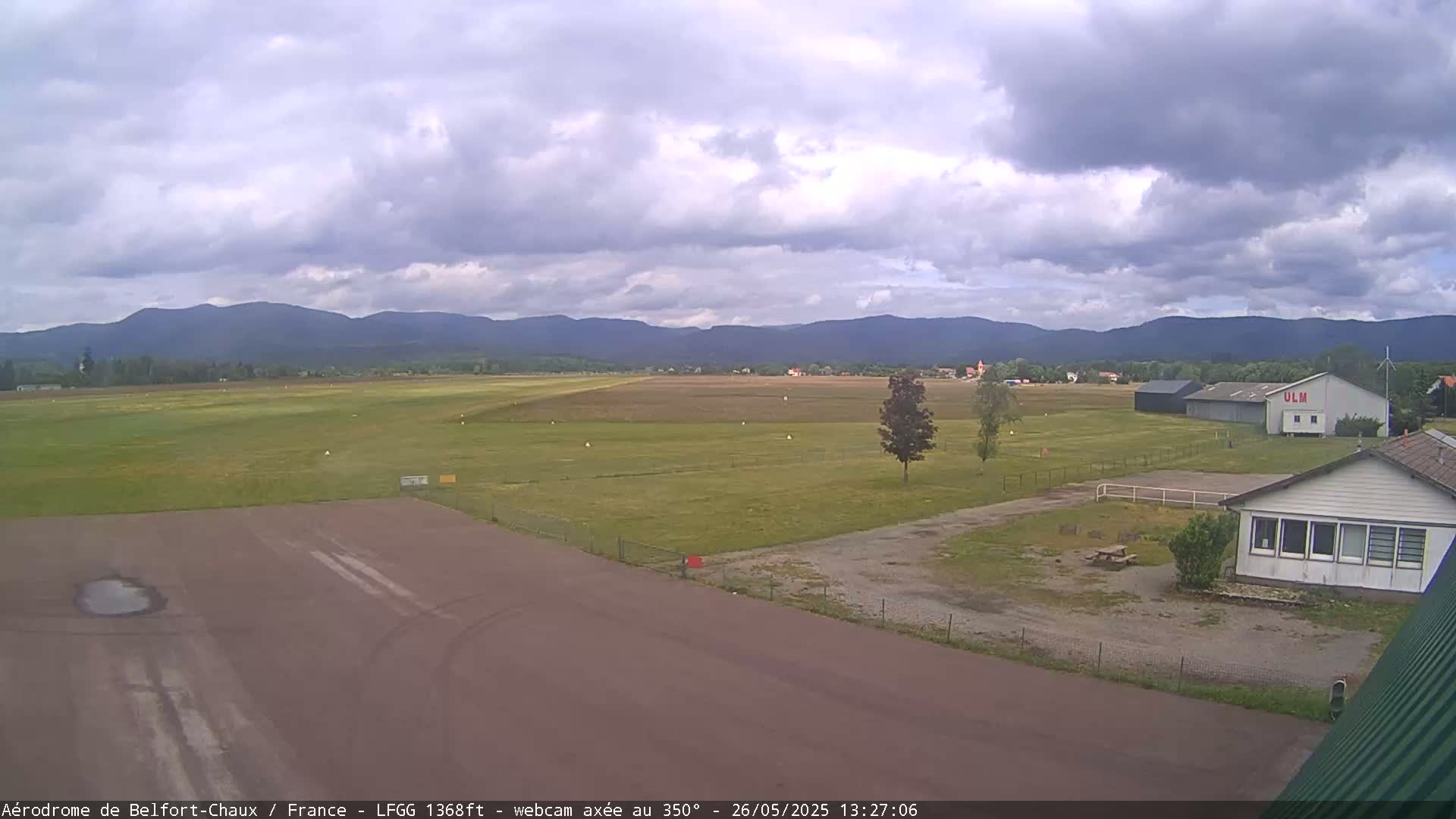 A mostly cloudy day at an airfield shows a runway, a small building, and distant mountains.