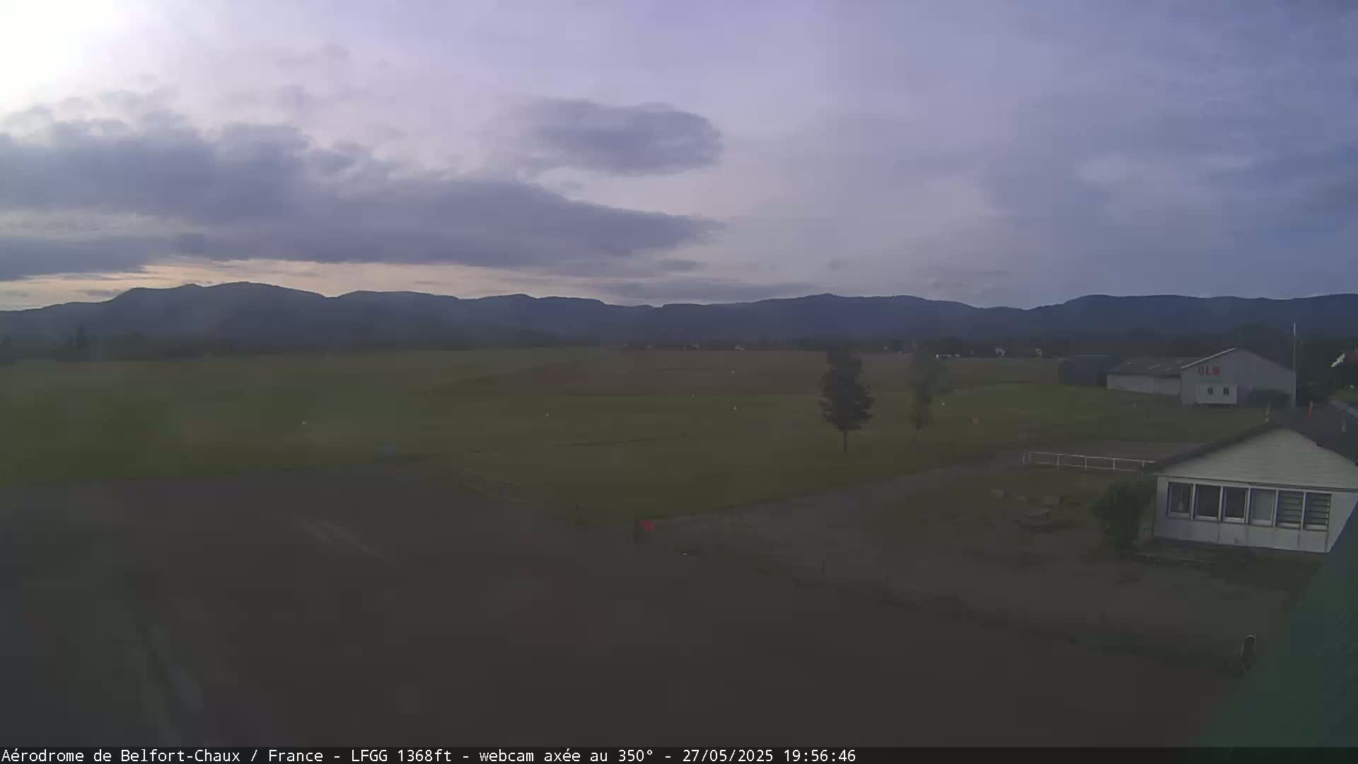 A mostly cloudy, twilight scene shows a flat green field with a few buildings and distant mountains.