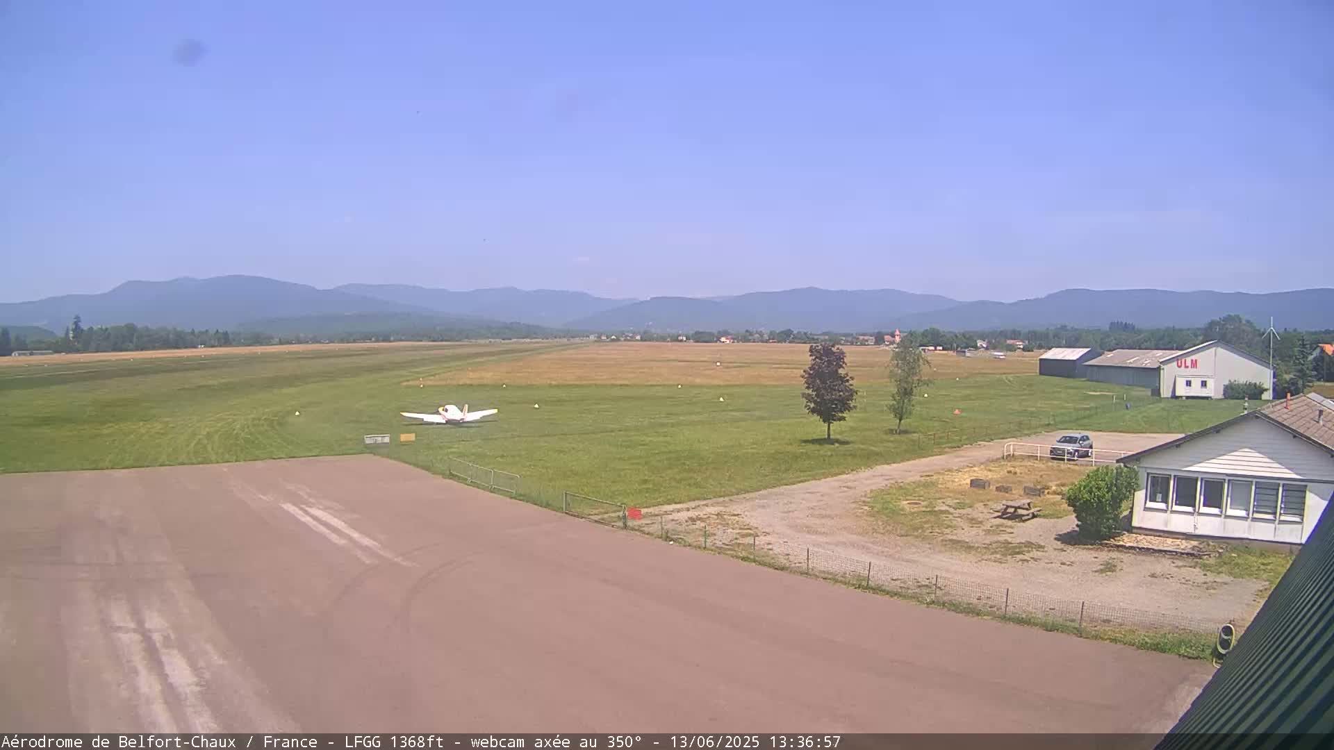 A small airplane sits on a grassy airfield under a clear blue sky, with distant mountains visible in the background.