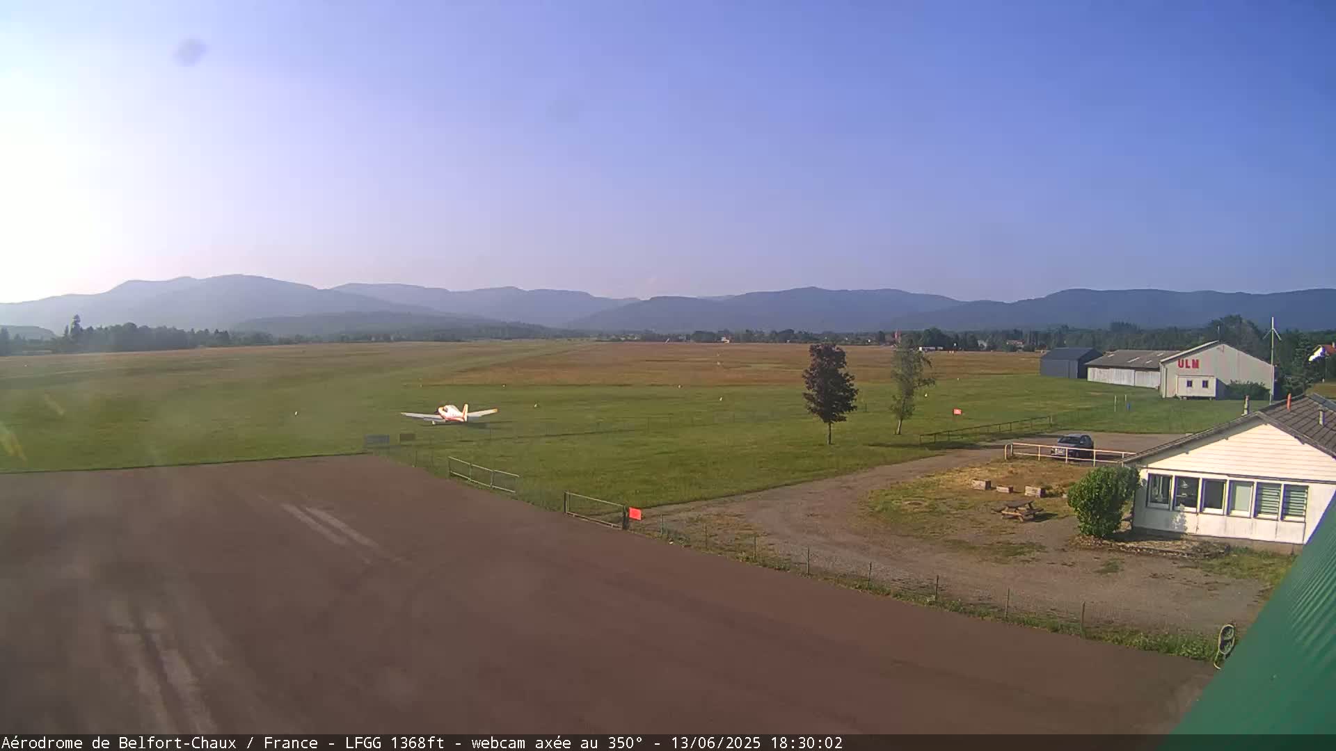 A small airplane sits on a grassy airfield, with a hangar and buildings visible in the foreground, and a mountain range under a clear blue sky in the background.
