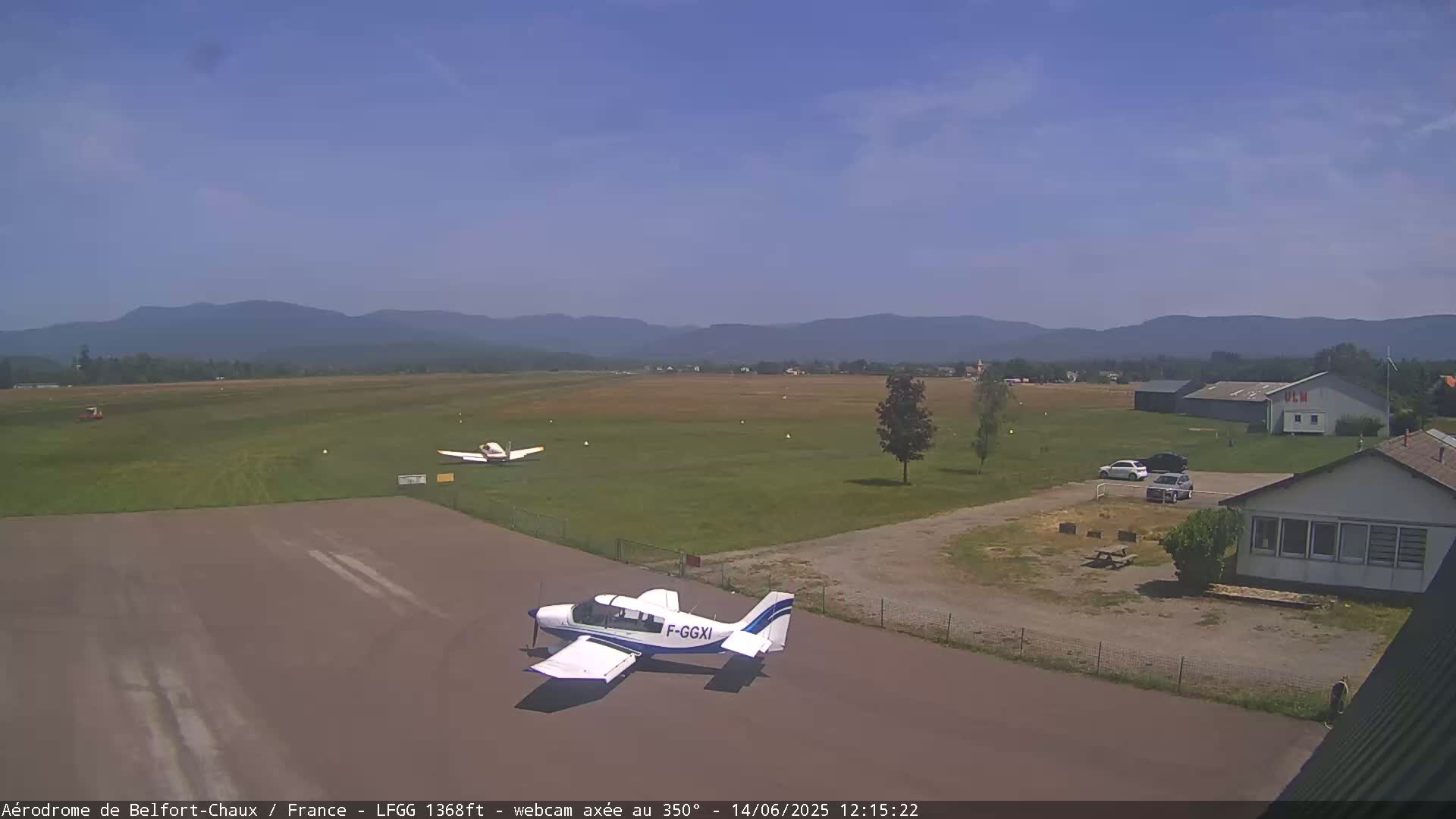 A small airfield with two light aircraft on the tarmac, a few cars parked nearby, and a building in the background under a clear blue sky.