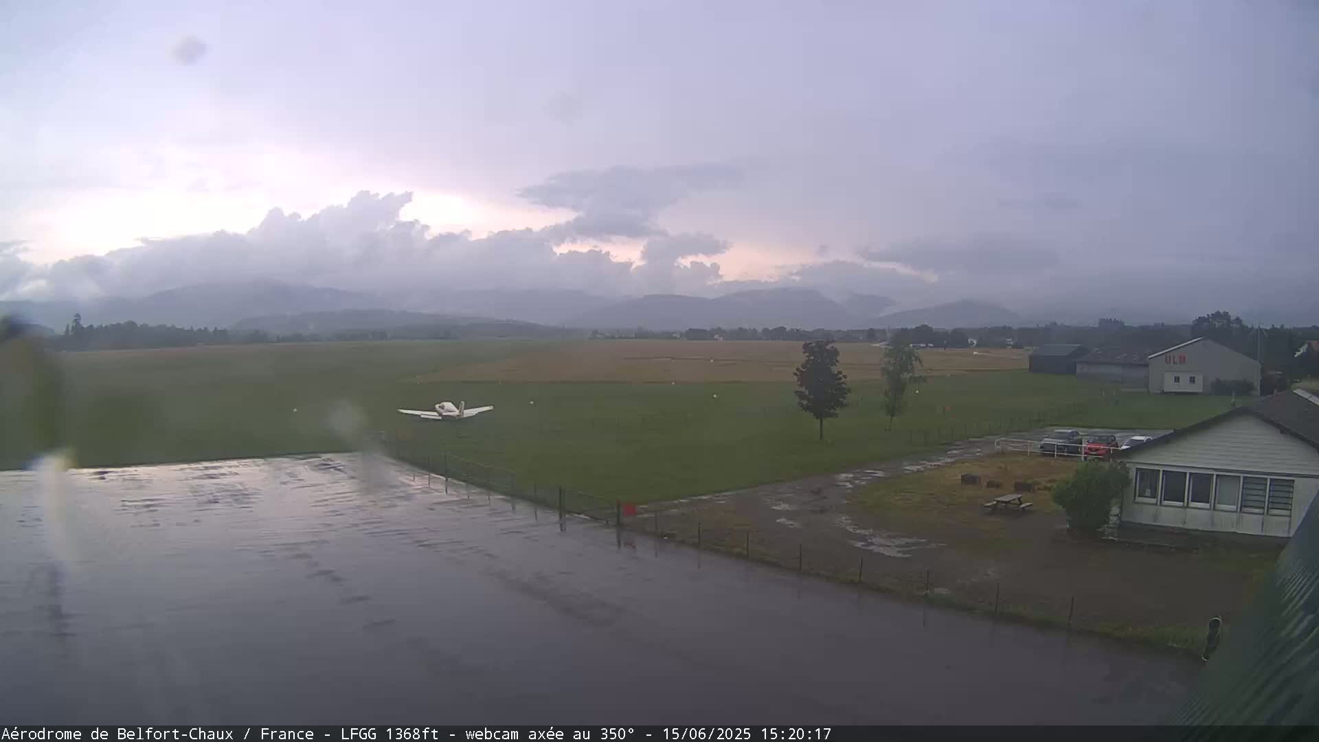 A small airplane sits on a rain-slicked airfield before a line of distant mountains under an overcast sky.