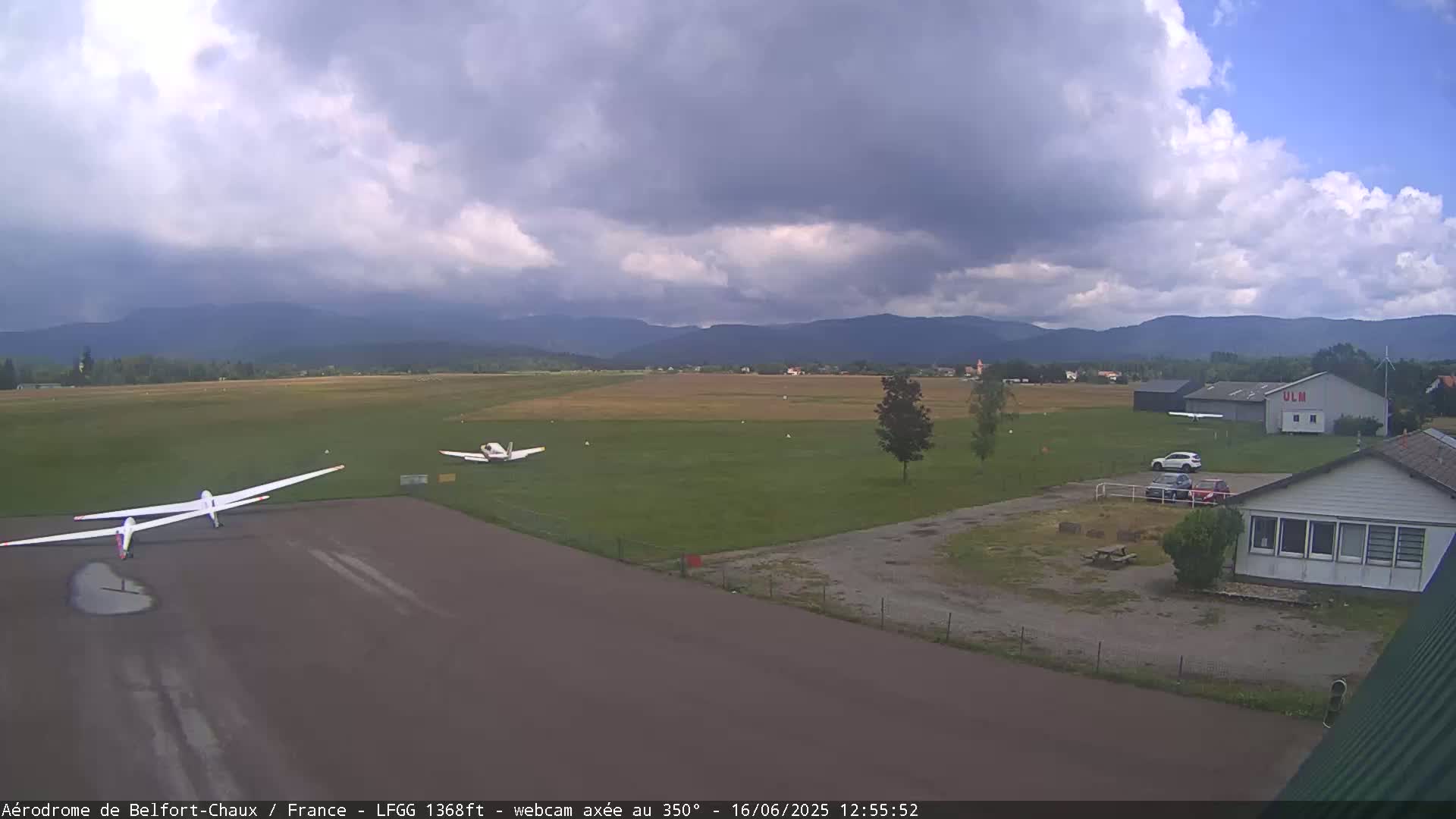 Two gliders and a small airplane are on a grassy airfield under a partly cloudy sky with mountains in the background.