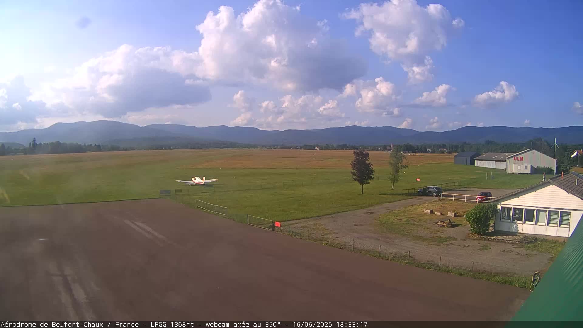 A small airplane sits on a grassy airfield under a partly cloudy sky, with mountains visible in the distance.