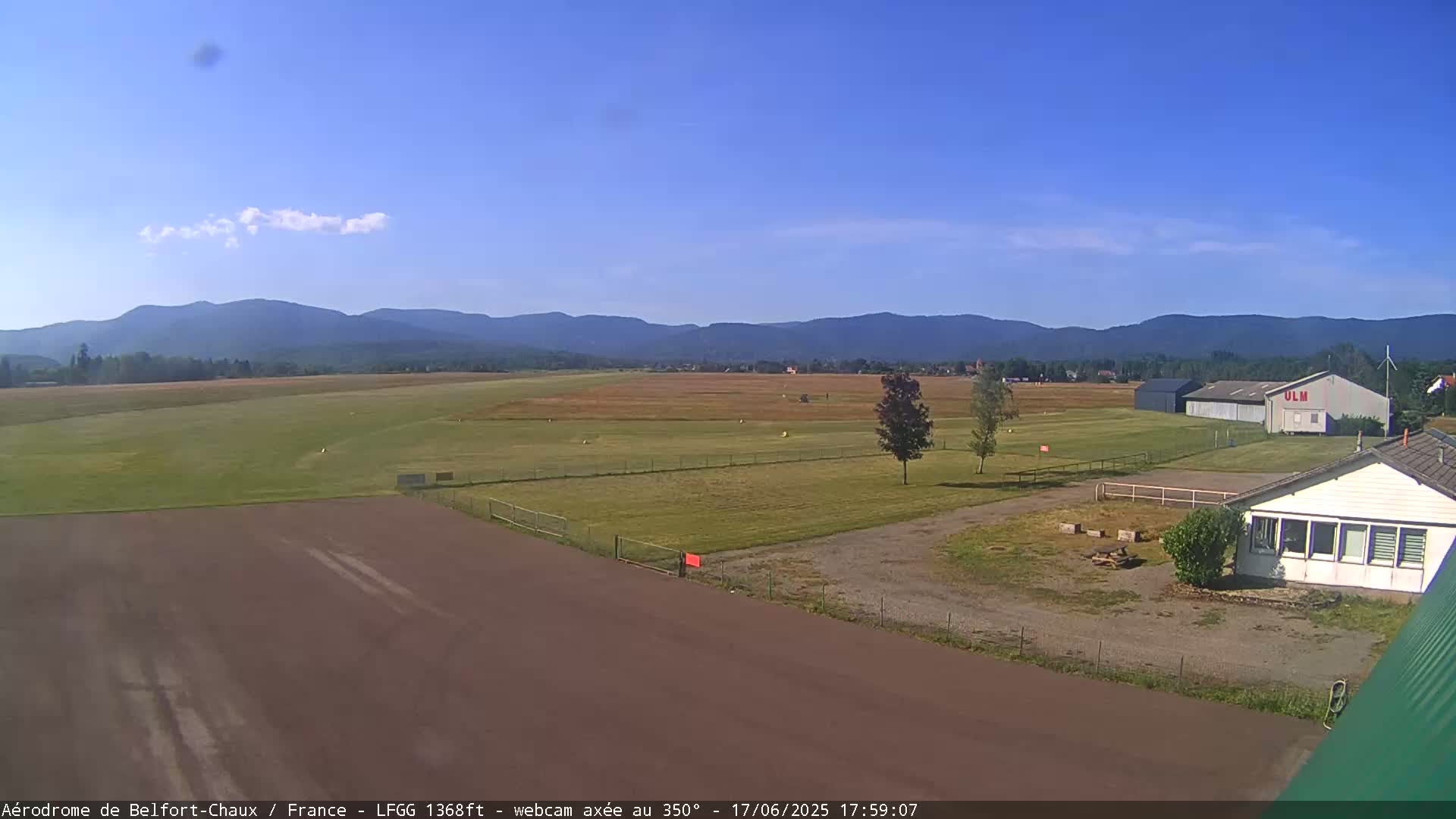 A small airfield with a few buildings is situated in a grassy field under a clear blue sky, with mountains visible in the distance.