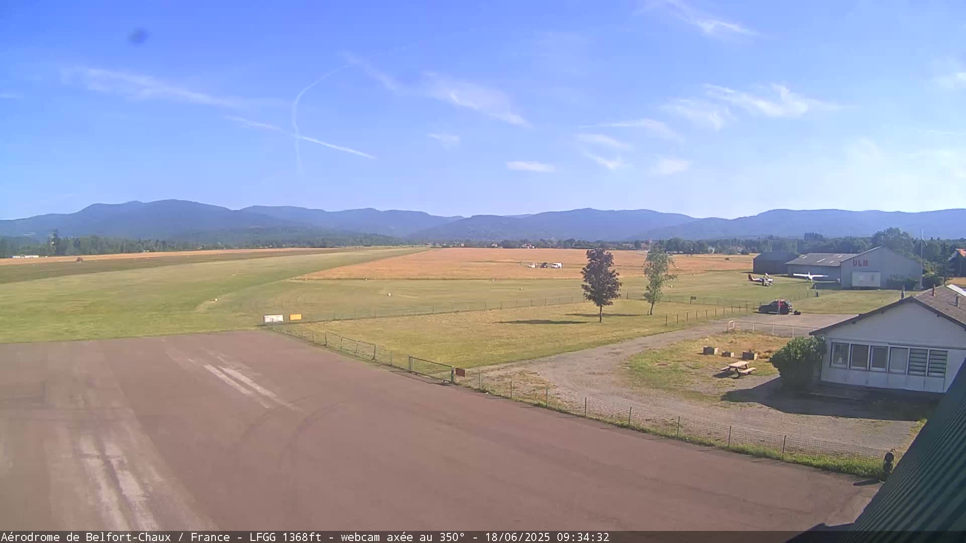 A small airfield with several small aircraft, a hangar, and a small building is situated in a grassy field, with mountains visible in the background under a mostly clear sky.