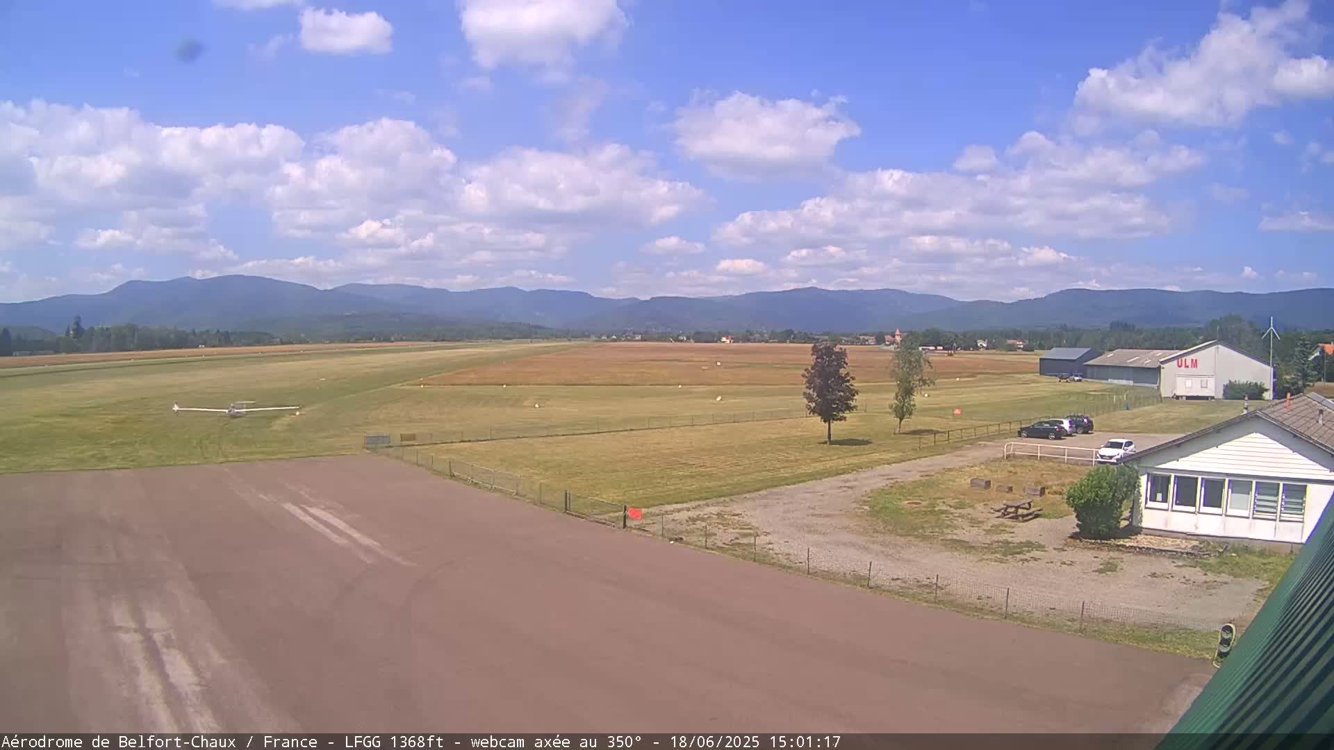 A small airfield with a glider on the runway, a few cars, and buildings, is situated under a partly cloudy sky with distant mountains in the background.