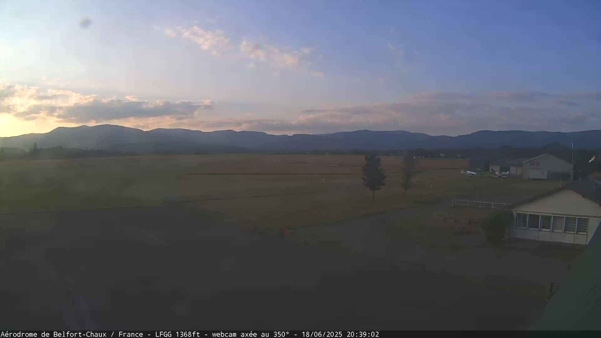 A flat, grassy airfield with a few small planes and buildings is visible under a mostly clear, twilight sky, with distant mountains in the background.