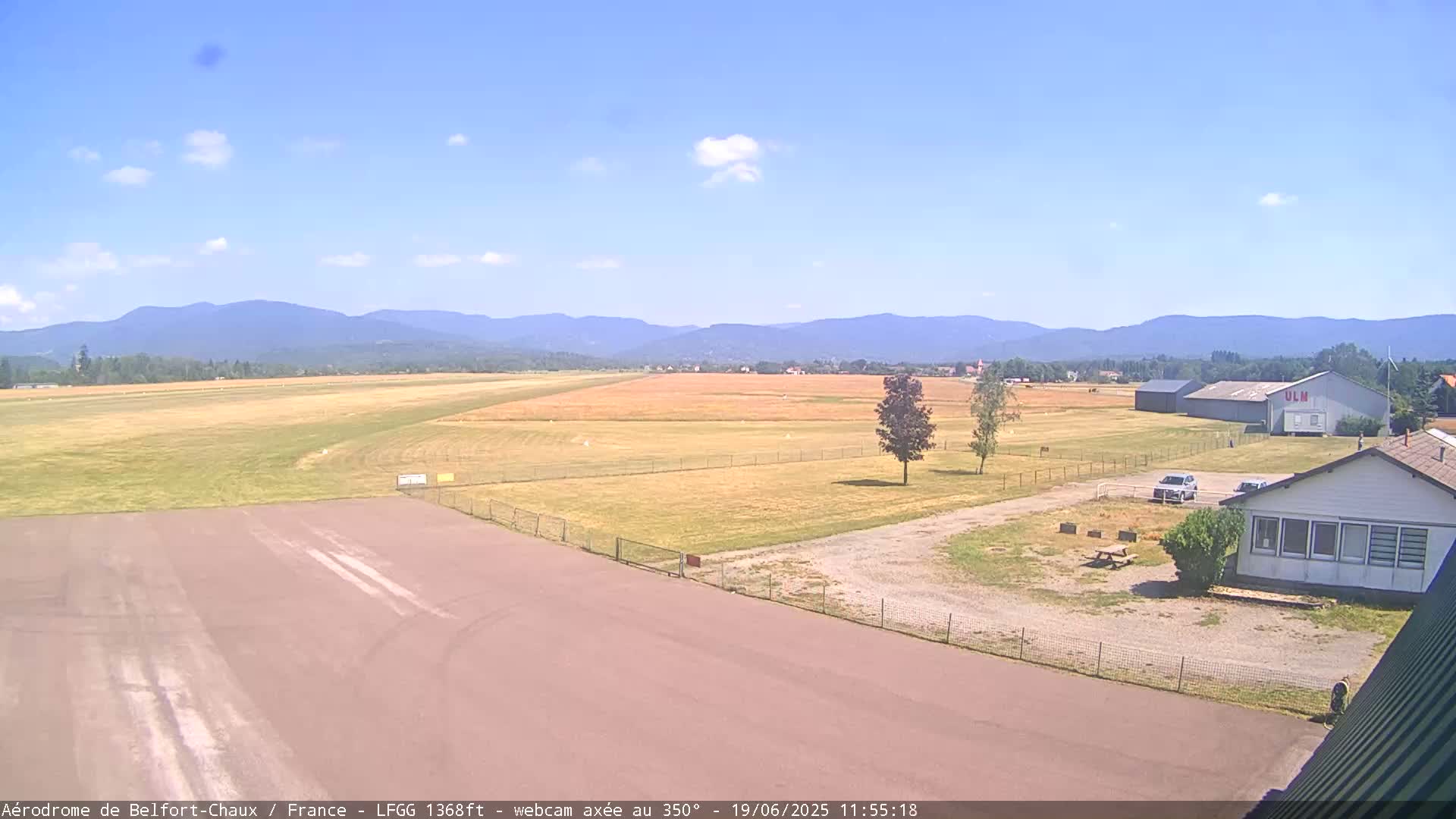 A small airfield with a runway, grassy fields, and a few buildings is visible under a mostly sunny sky with a few scattered clouds, and mountains are seen in the distance.
