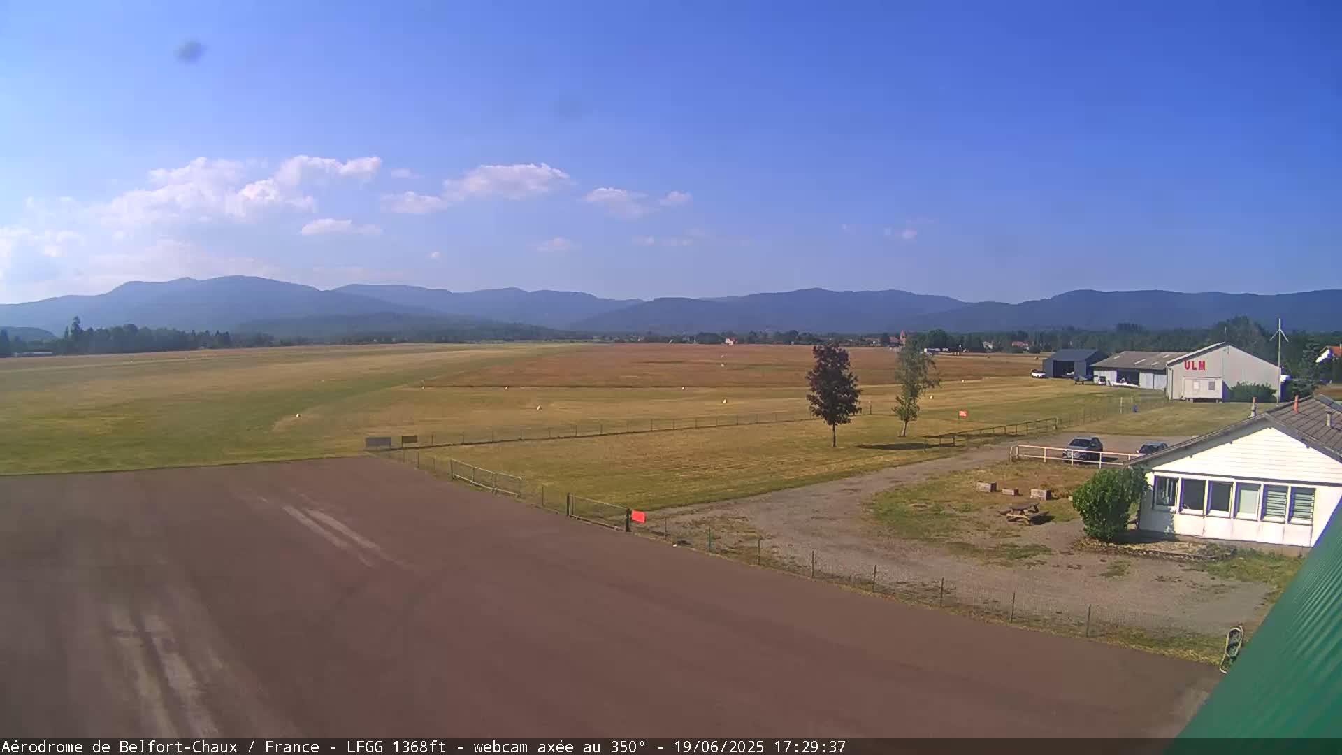A small airfield with a hangar and a building is situated in a grassy field, backed by a range of distant mountains under a mostly clear, bright sky.