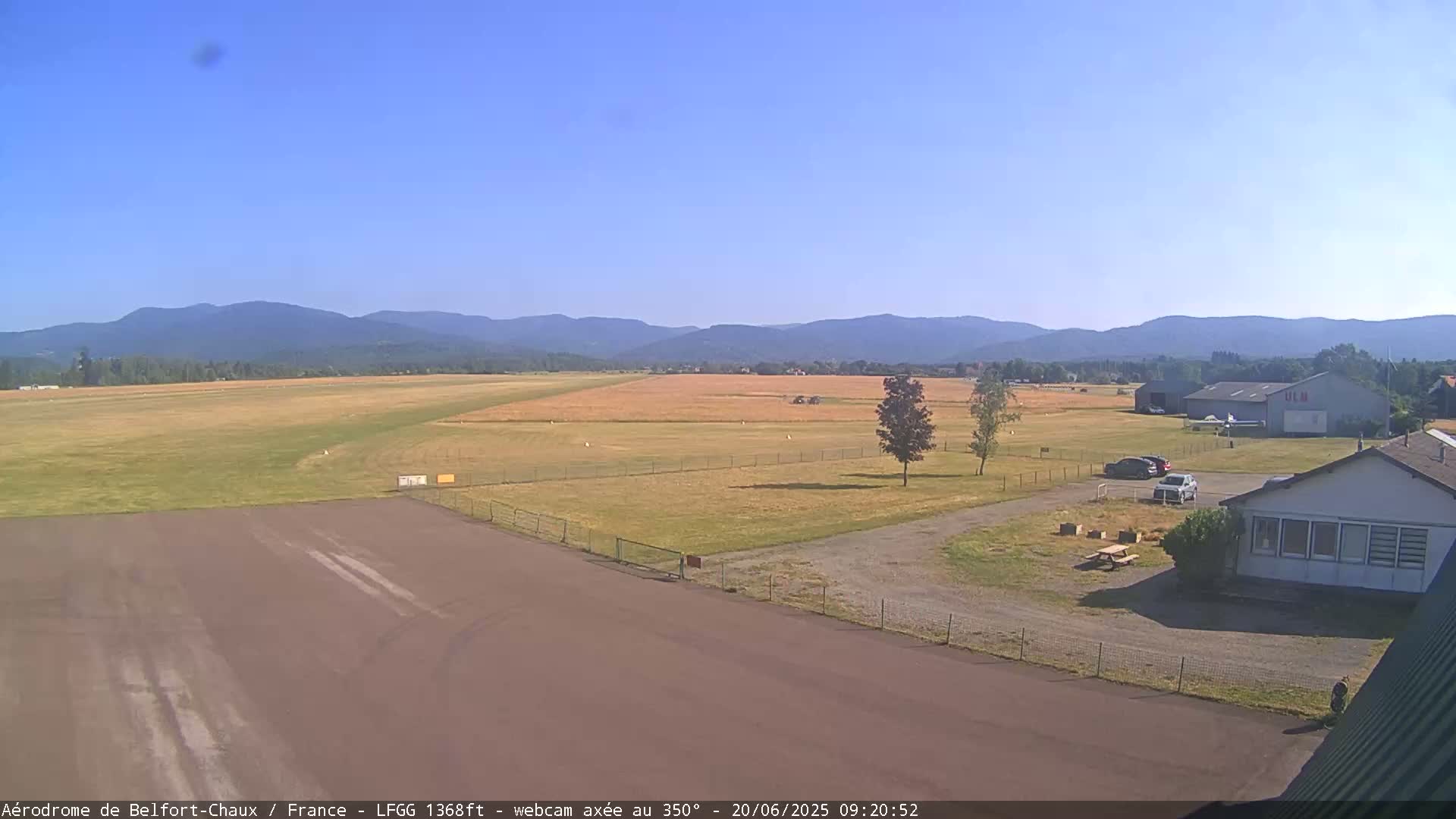 A paved airfield runway extends to a grassy field, bordered by a few buildings and trees, under a clear blue sky and mountains in the distance.