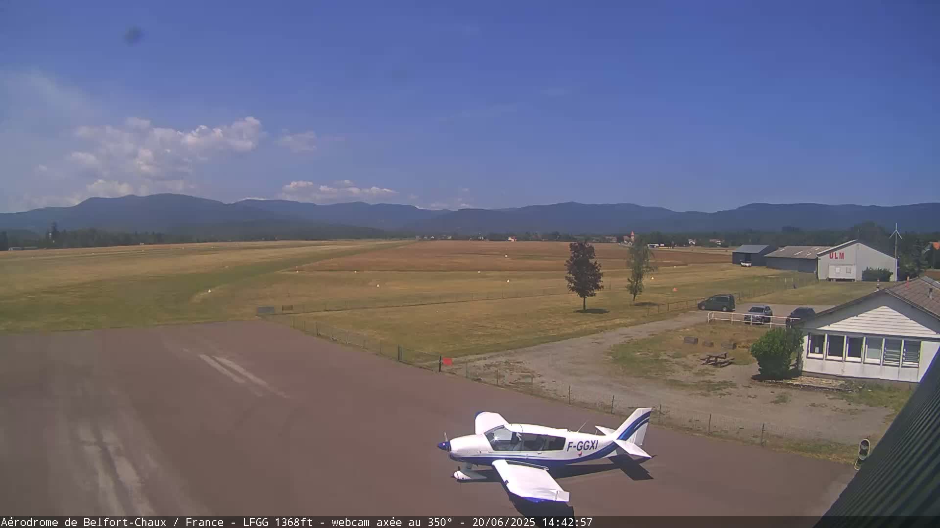 A small airplane sits on a tarmac at a small airport on a partly cloudy day with mountains in the background.