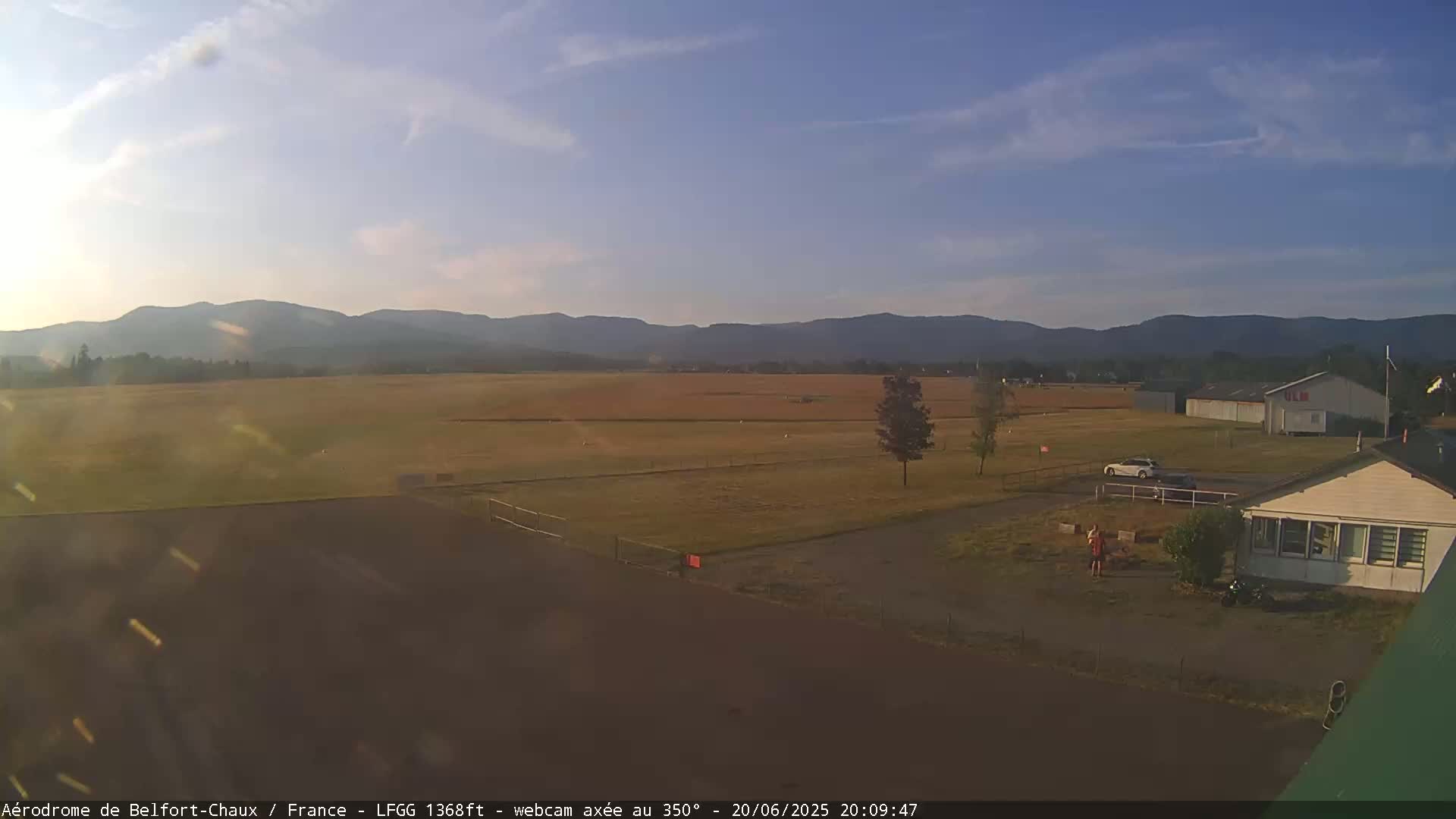 A flat, grassy airfield with a few buildings and a car is visible under a clear, mostly sunny sky with distant mountains in the background.