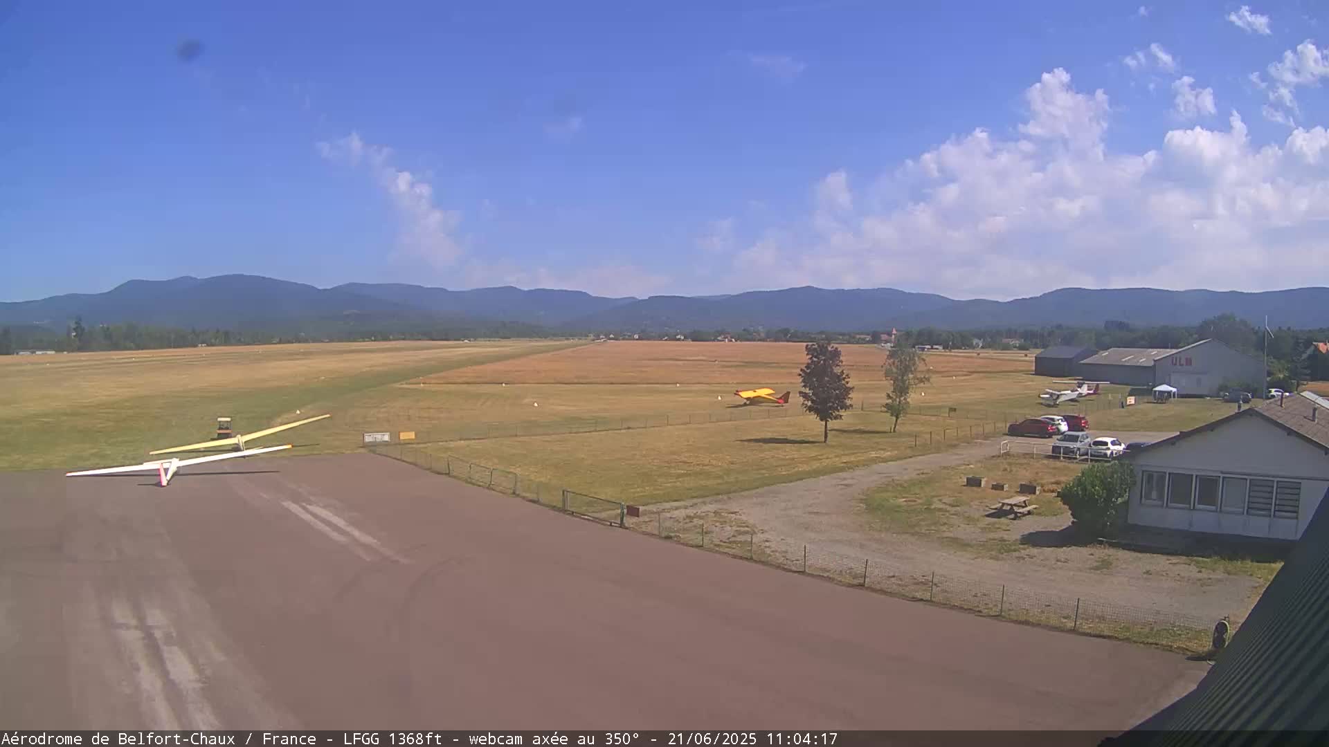 A small airfield with several small airplanes and gliders on a sunny day with partly cloudy skies and mountains in the background.
