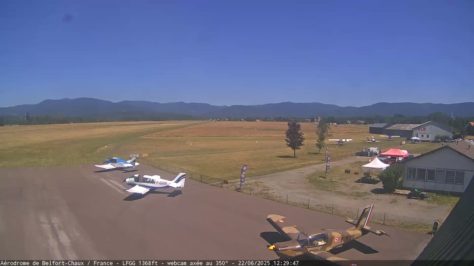 A small airport with several small airplanes on the tarmac, under a clear blue sky and in front of a mountain range.