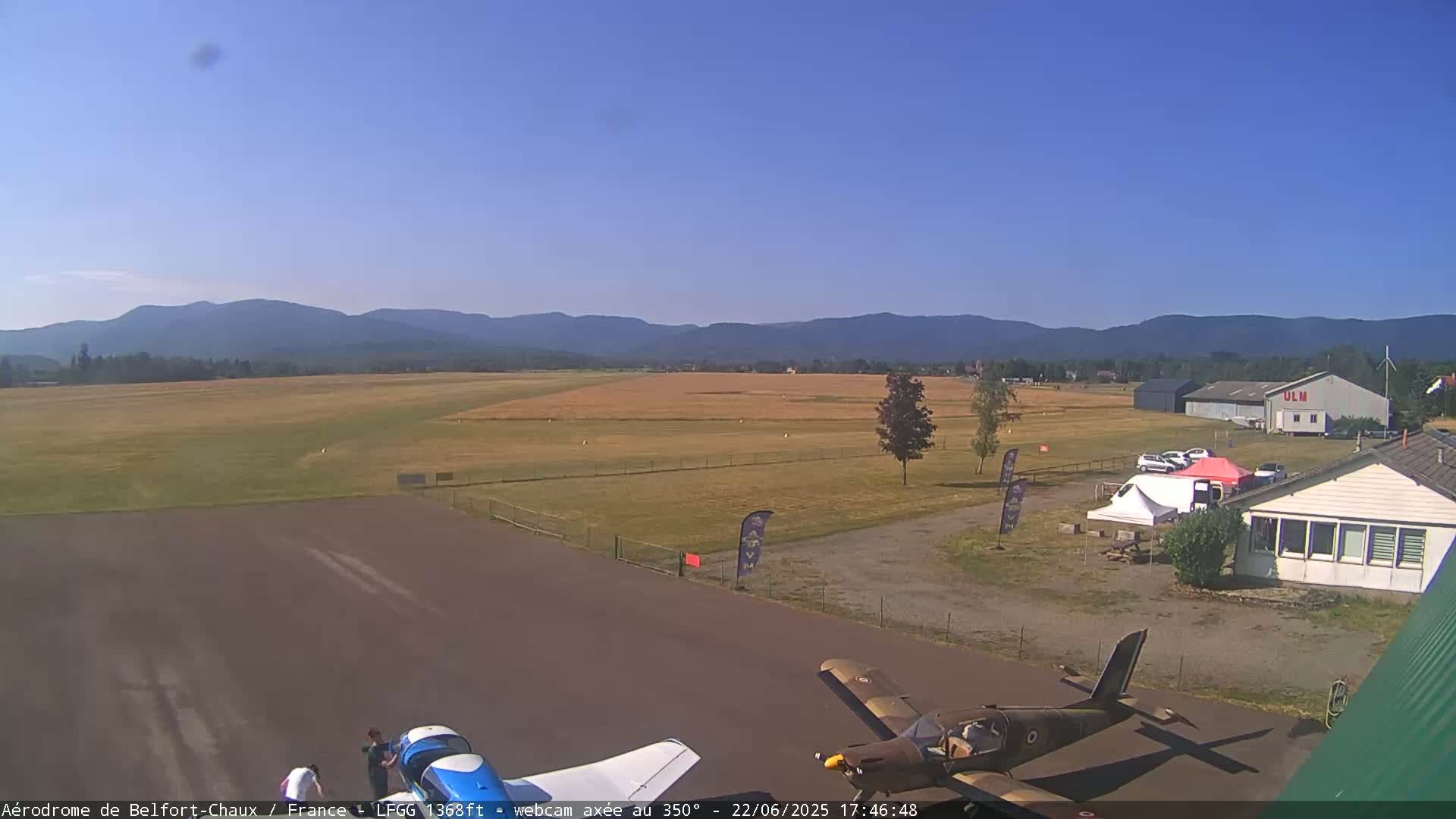 A small airport with two airplanes on the tarmac, several buildings, and tents under a clear blue sky with distant mountains in the background.