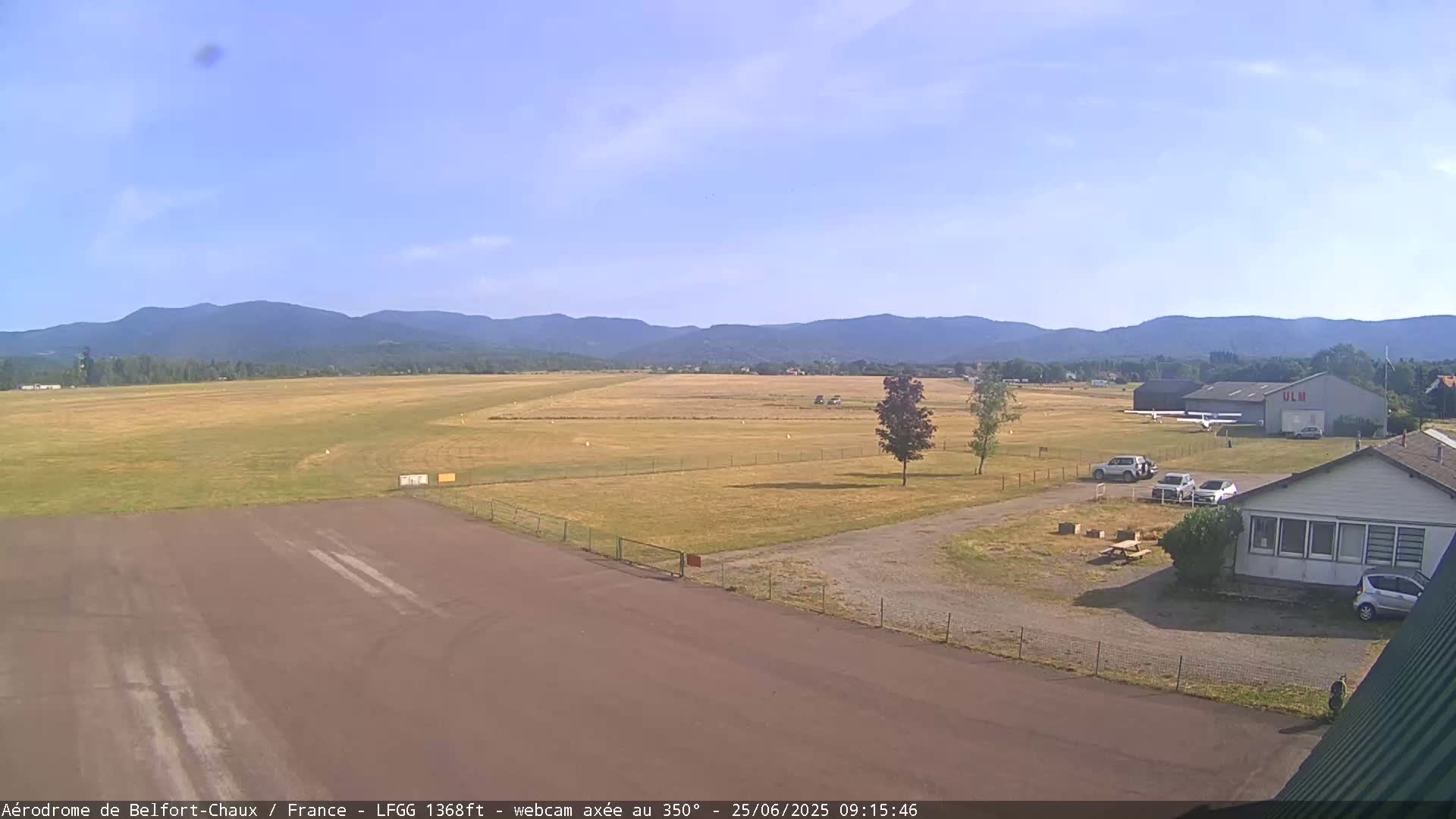 A small airfield with a hangar and several cars parked nearby, situated in front of a grassy runway and backed by a range of mountains under a clear sky.