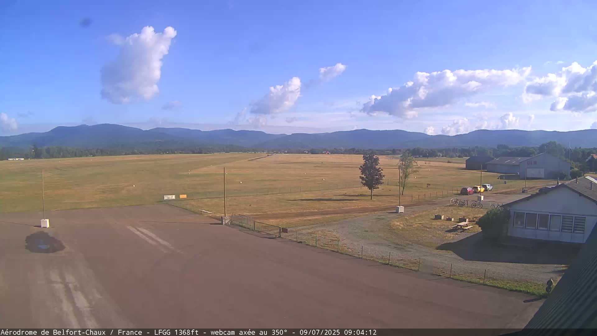 A mostly sunny day at an airfield shows a runway, a few buildings, and distant mountains under a partly cloudy sky.
