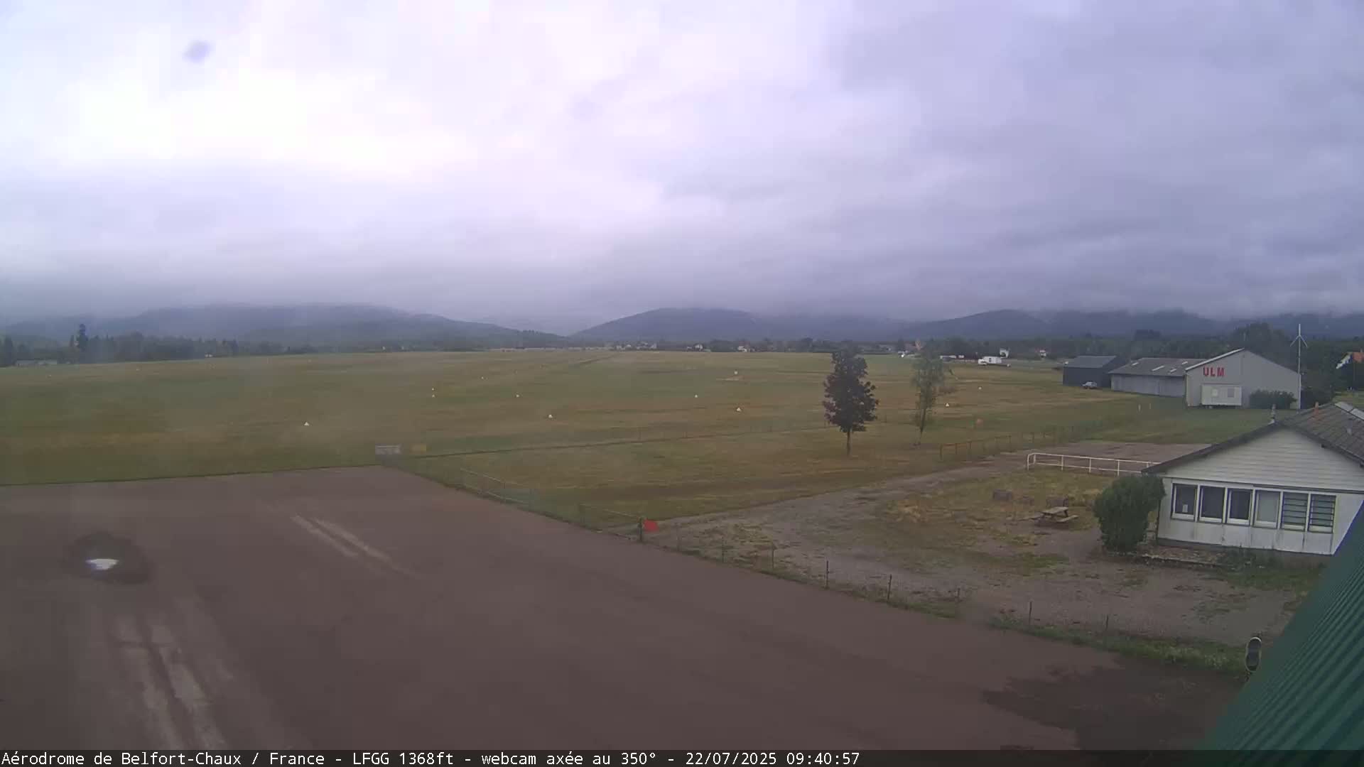 A mostly overcast sky hangs over a grassy airfield with a few buildings, including a hangar, and distant mountains.