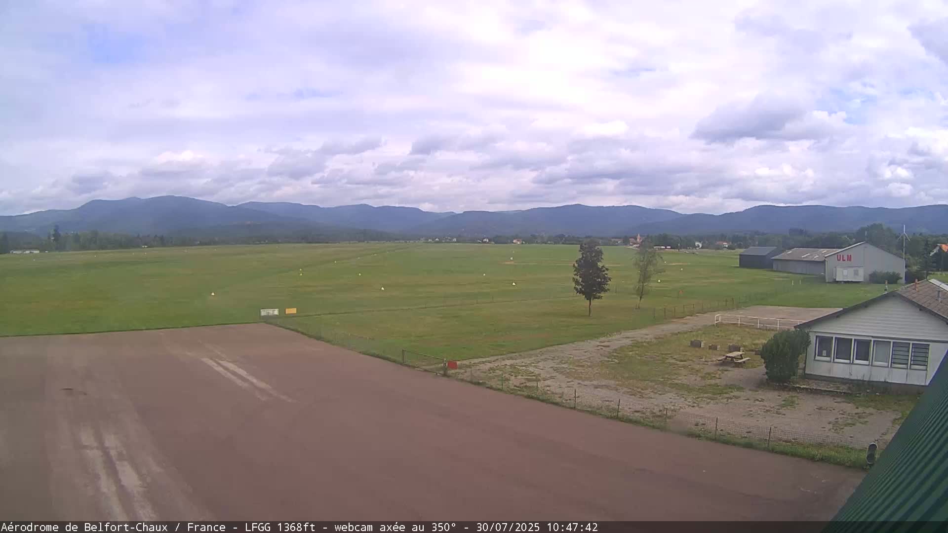 A mostly clear day shows a grassy airfield with a runway, small buildings, and distant mountains.