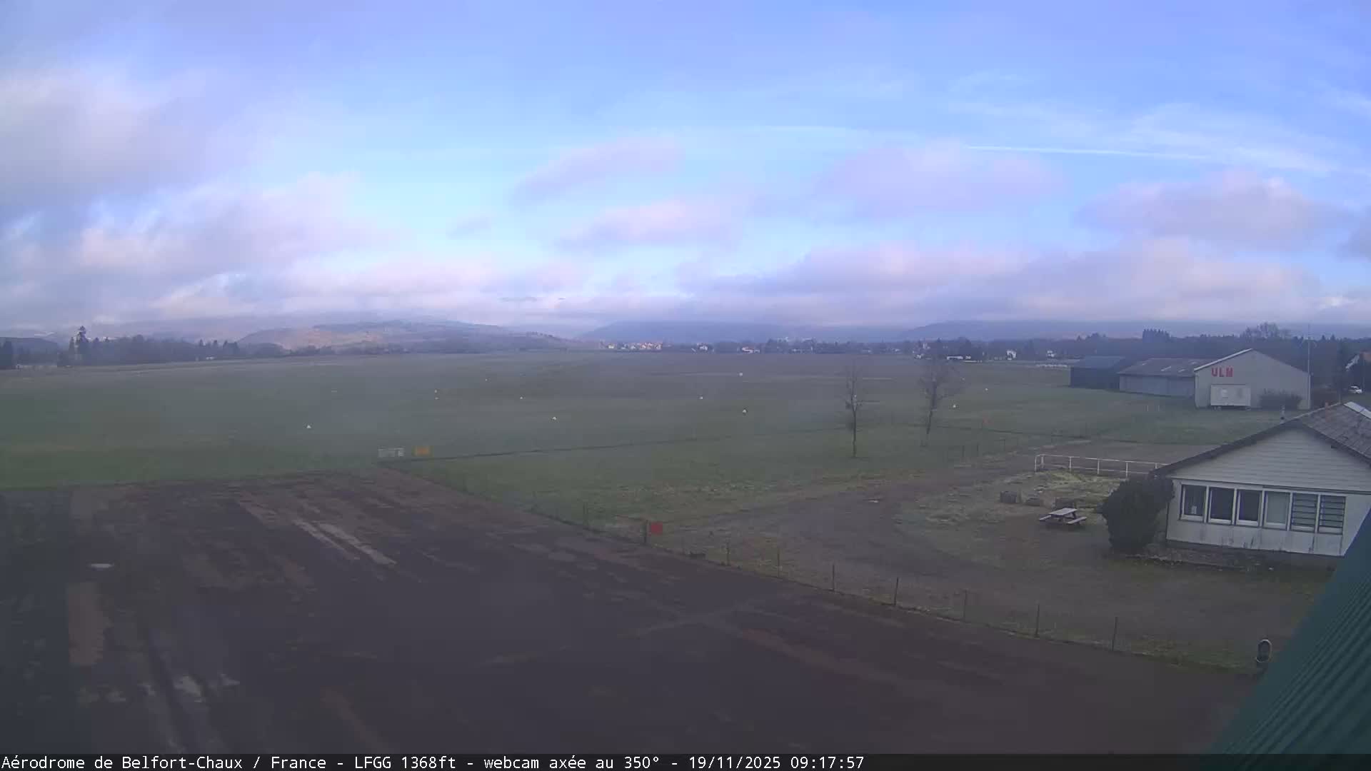 The image shows an airfield with a large grassy field, a dark runway in the foreground, and several buildings on the right, all under a sky with scattered clouds and patches of blue, suggesting cool, damp weather with hazy distant hills.