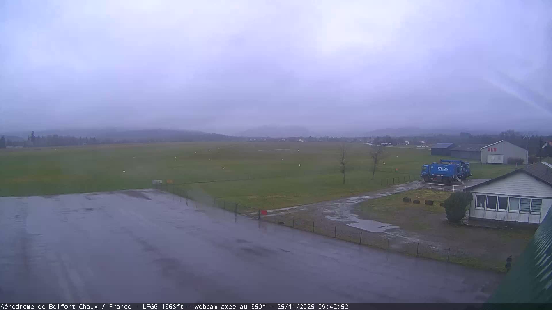 A wide, elevated view reveals a wet airfield on an overcast day, featuring a vast green runway, damp paved areas, several buildings, two blue service trucks, and distant hills obscured by mist under a uniformly grey sky.