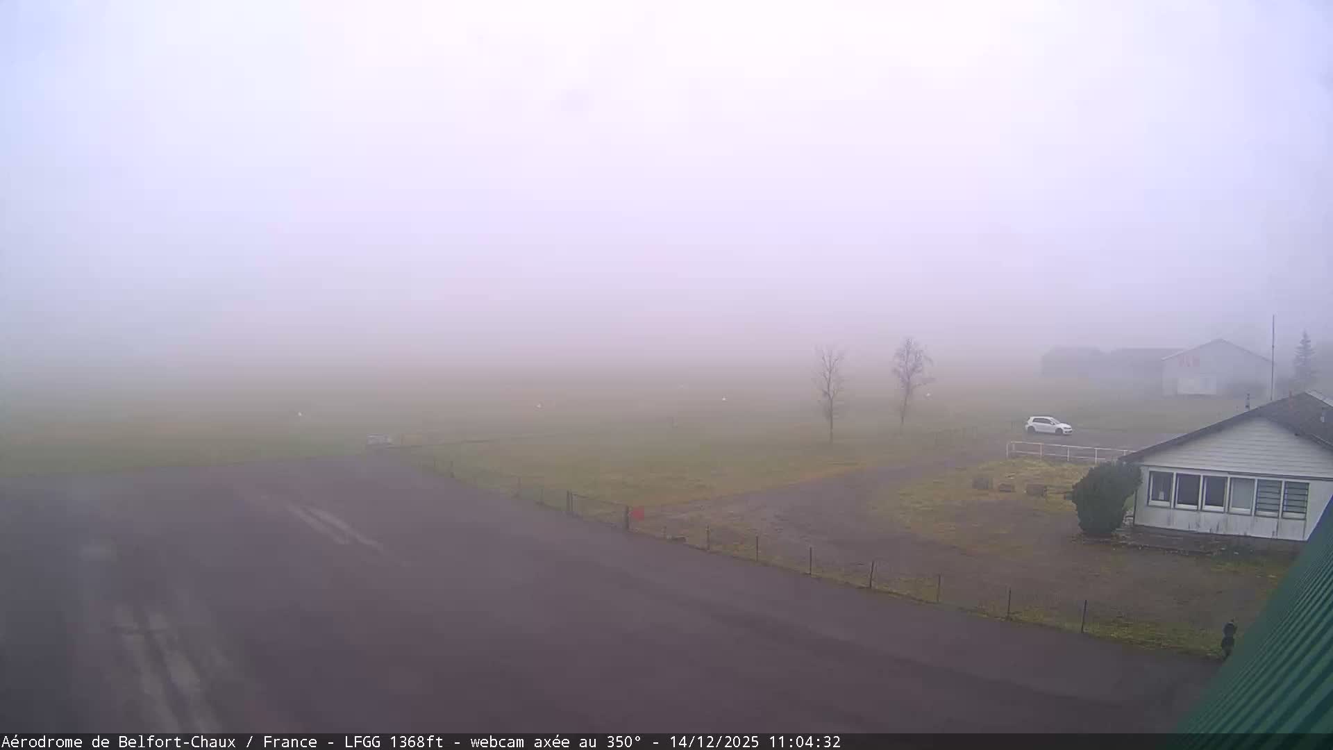 A wide, elevated view reveals a wet airfield on an overcast day, featuring a vast green runway, damp paved areas, several buildings, two blue service trucks, and distant hills obscured by mist under a uniformly grey sky.