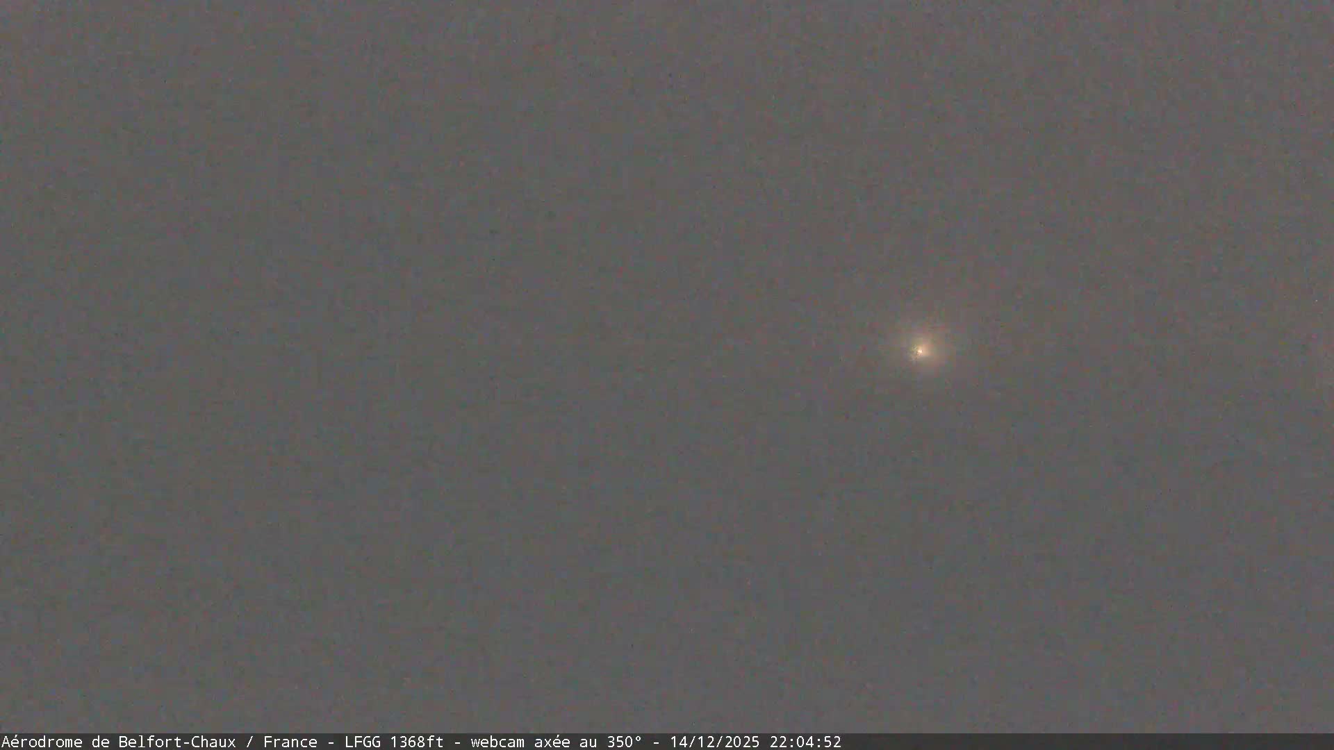A wide, elevated view reveals a wet airfield on an overcast day, featuring a vast green runway, damp paved areas, several buildings, two blue service trucks, and distant hills obscured by mist under a uniformly grey sky.