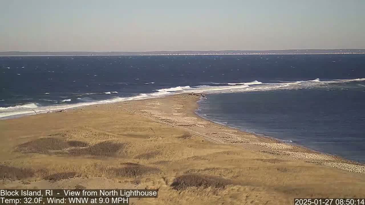A sandy beach curves into a bay of a windy, dark blue ocean under a clear sky.