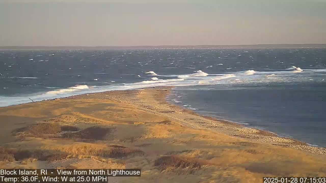 A sandy, dune-covered point of land juts into a choppy sea under a pale sky, with waves breaking along the shoreline.