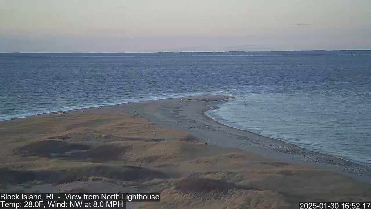 A sandy beach and a narrow spit of land extend into a calm, blue ocean under a clear, pale sky.