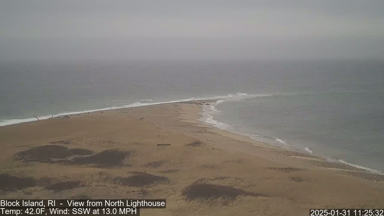 A hazy, overcast view shows a sandy beach and a narrow spit of land extending into a gray sea with small waves.