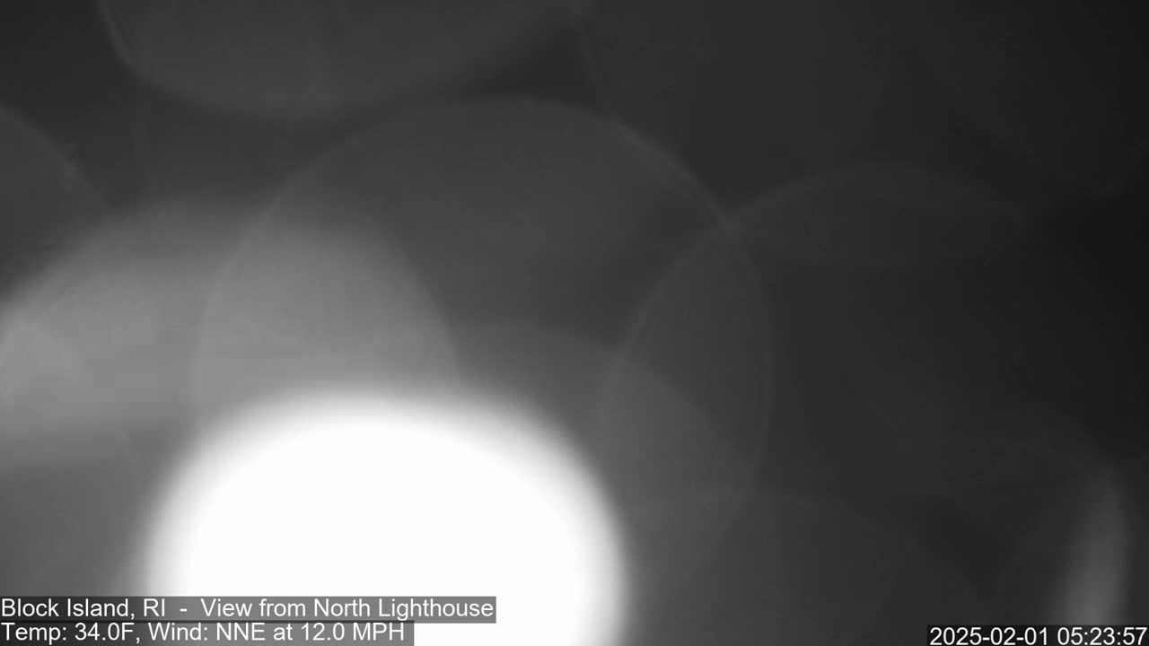 A blurry, grayscale image shows an out-of-focus view of a bright light source in the lower center against a dark background, taken in 34°F weather with a 12 mph wind from the north-northeast.