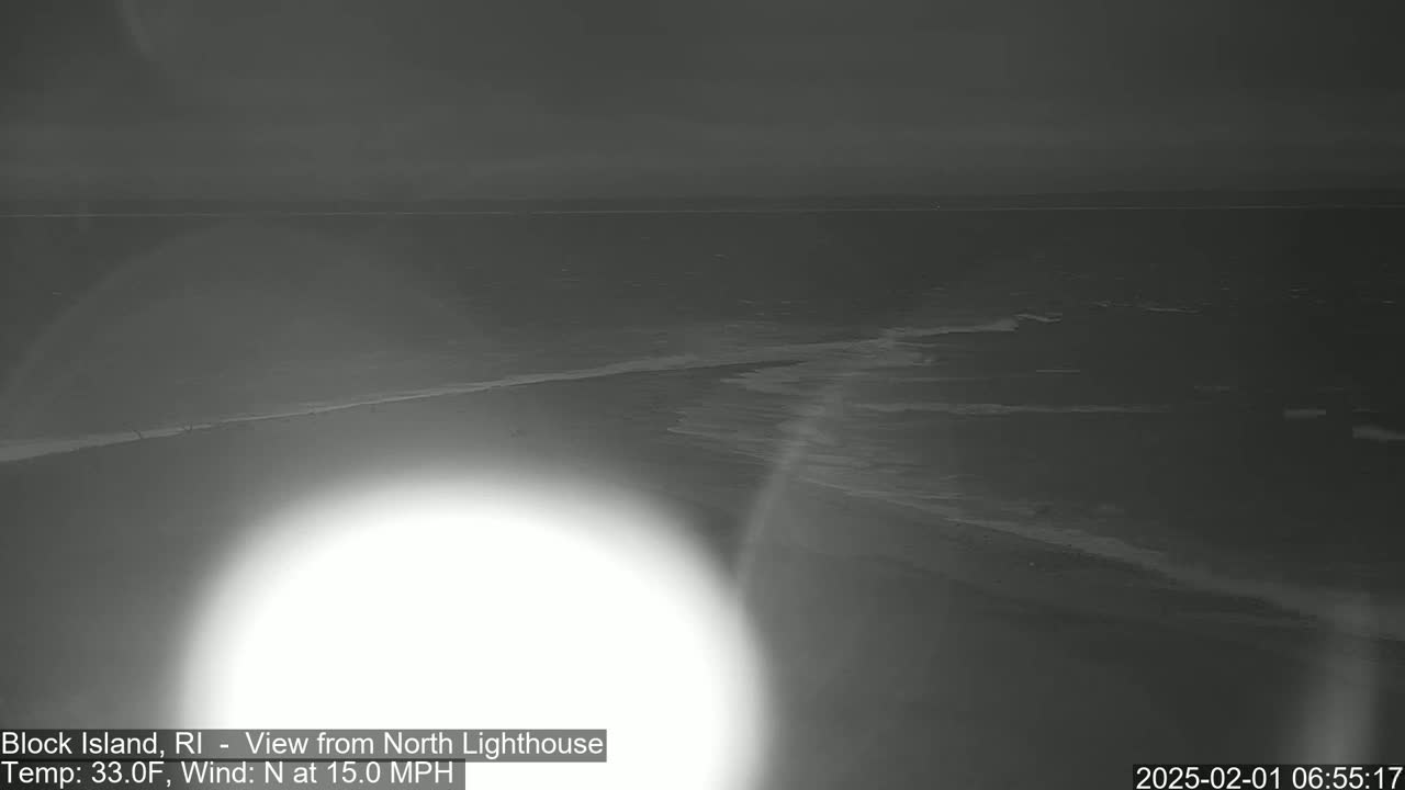 A nighttime coastal view shows a dark ocean with small waves under a gray sky, a bright light source obscuring part of the foreground.