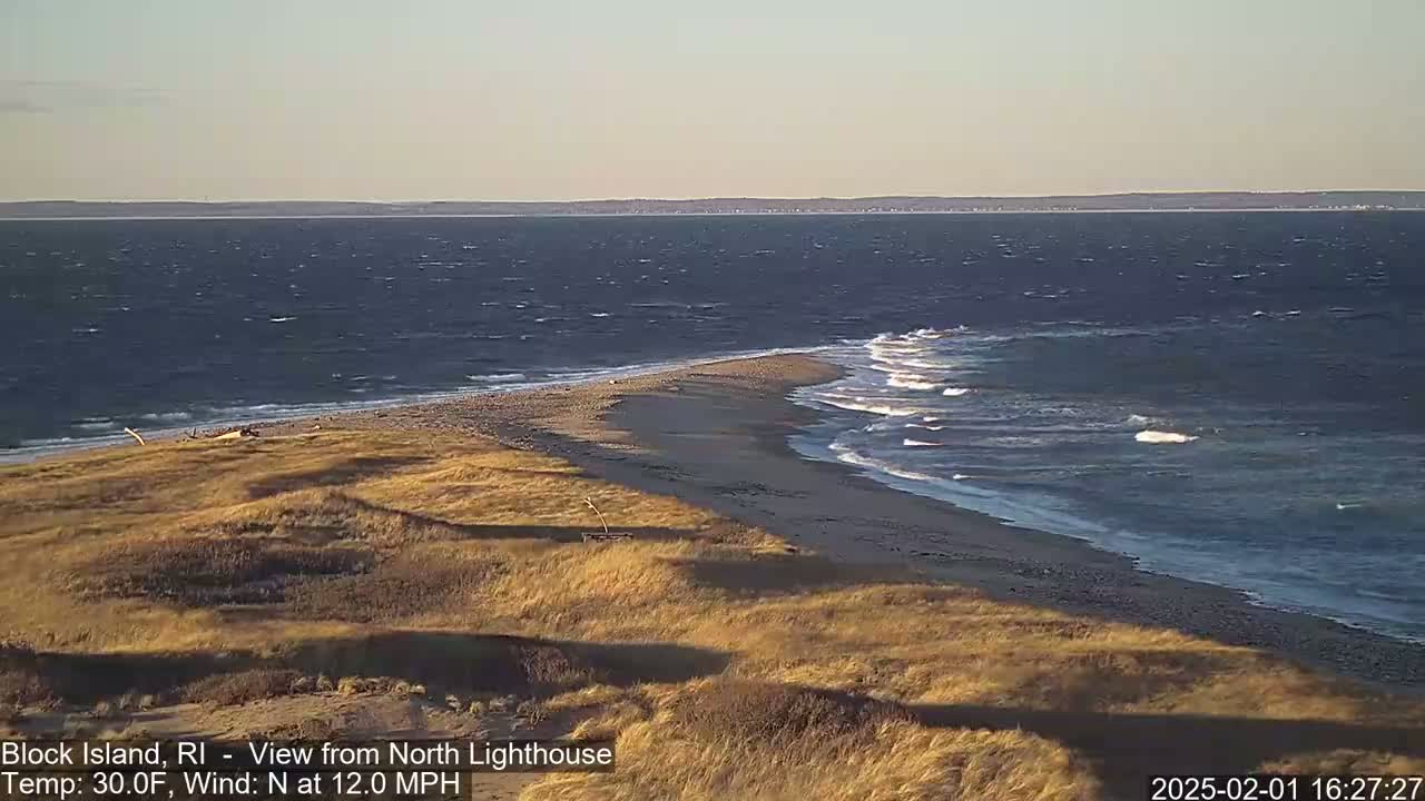 A sandy spit juts into a dark-blue ocean with moderate waves under a clear, pale sky.