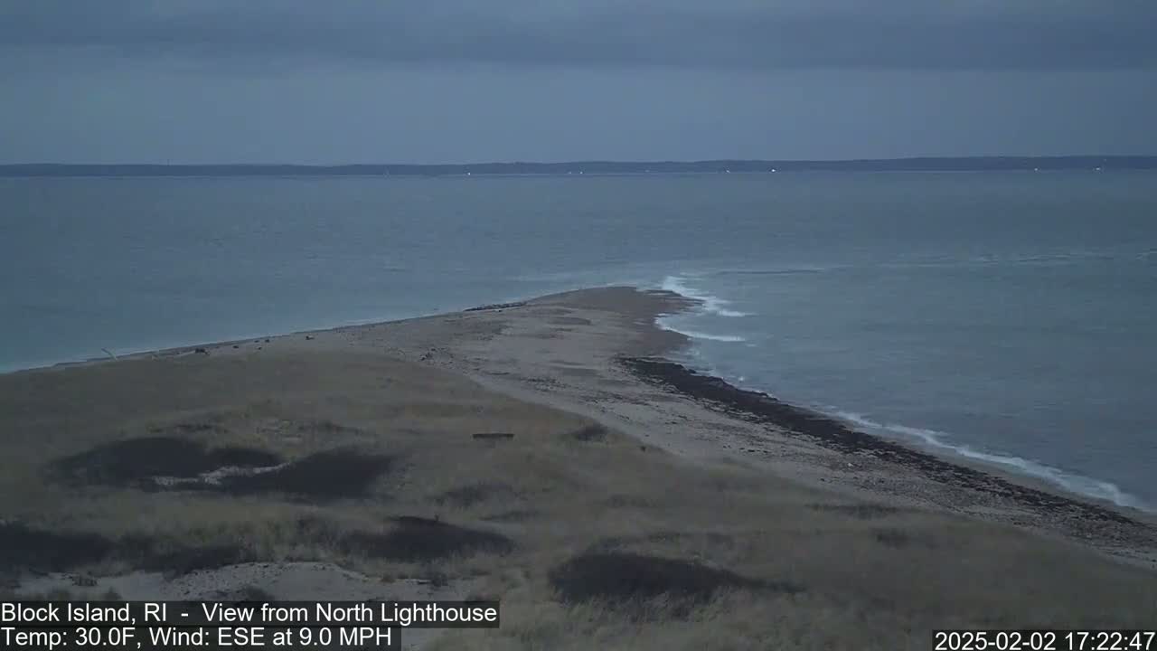 A sandy, low-lying spit of land juts into a calm, gray sea under a cloudy sky.