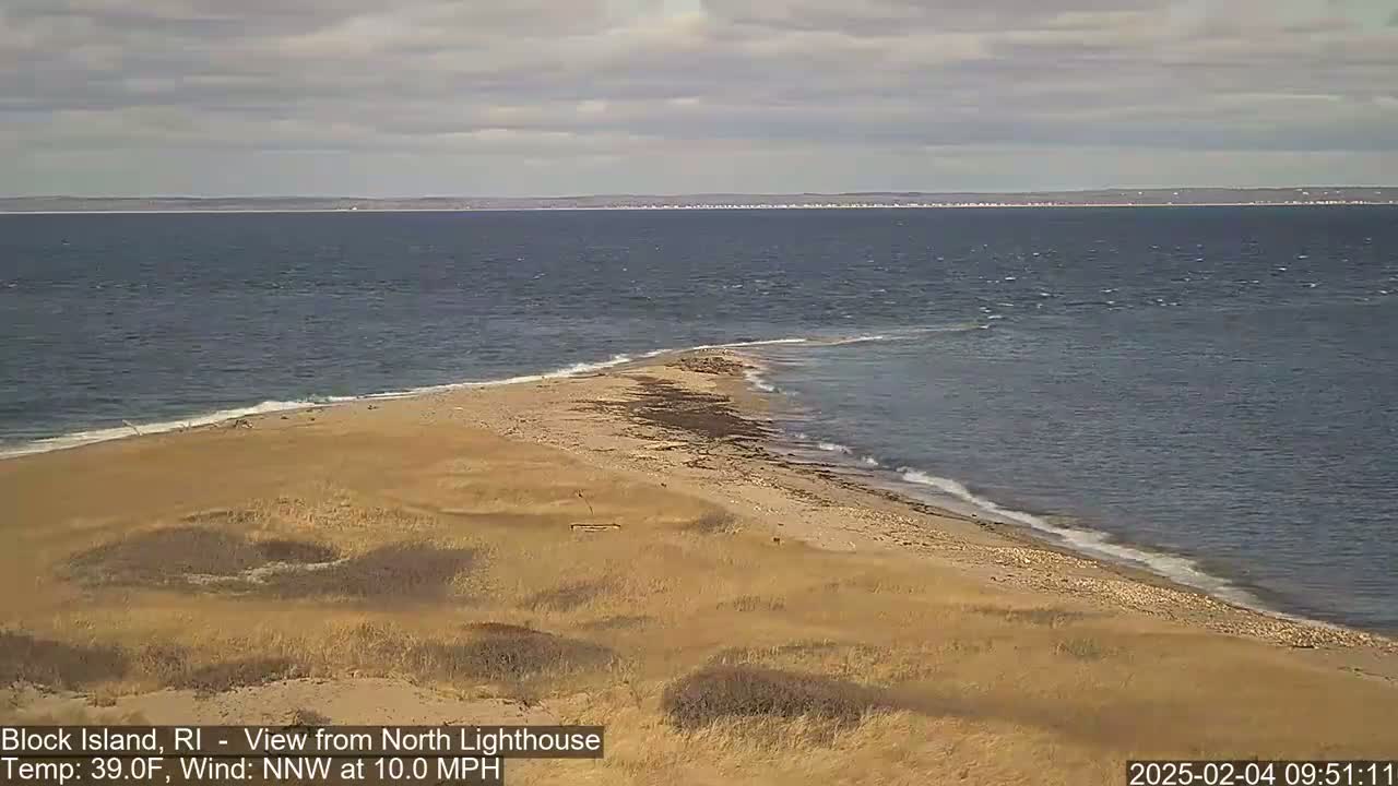 A sandy, grassy point of land juts into a calm, gray-blue ocean under a cloudy sky.