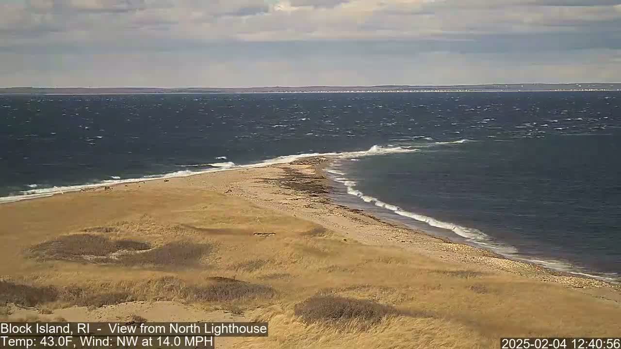 A sandy beach and a narrow spit of land jut into a choppy ocean under a partly cloudy sky.