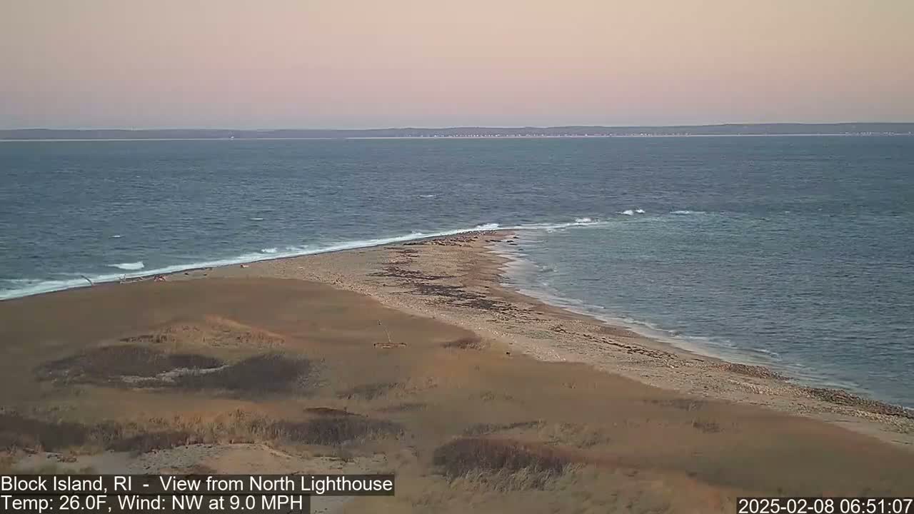 A sandy beach and ocean meet at a point of land under a pale pink sky with a northwest wind blowing at nine miles per hour.
