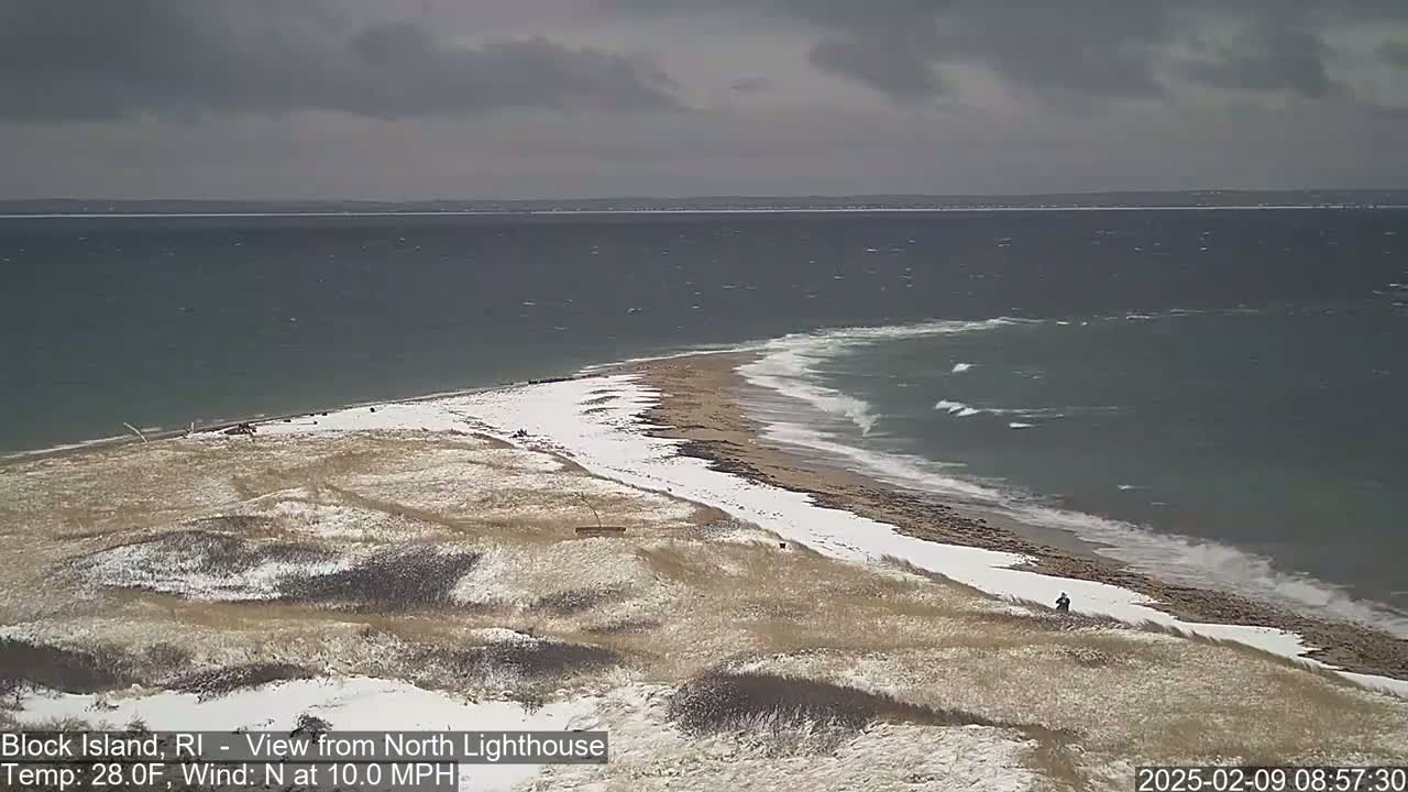 A snow-dusted beach and dune landscape meet a choppy ocean under a cloudy sky.