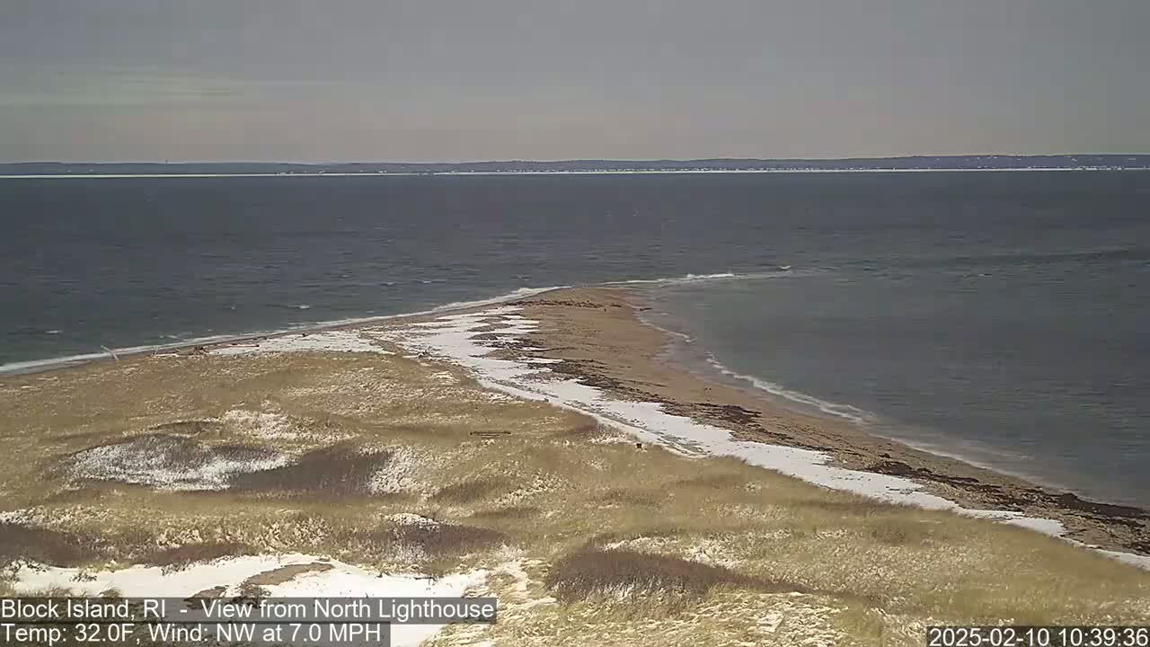 A snow-dusted sandy beach and dune landscape meet a calm, gray ocean under a cloudy sky.