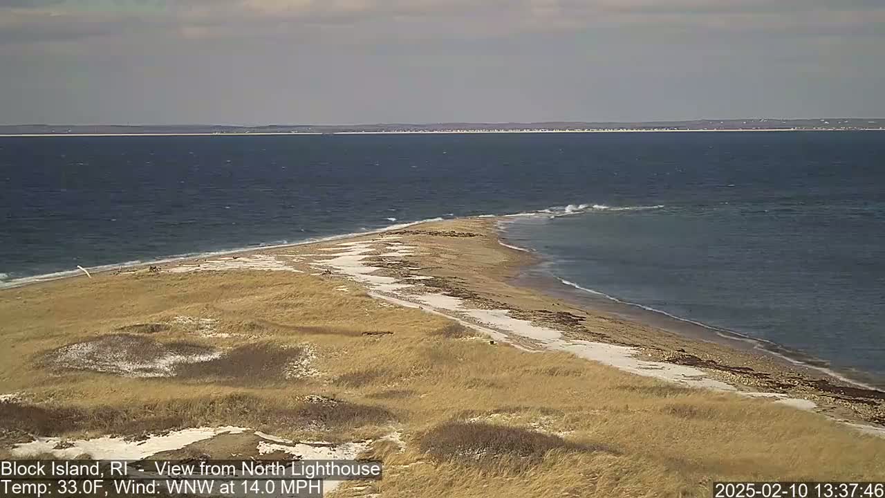 A sandy, grassy spit of land juts into a dark blue ocean under a mostly cloudy sky.