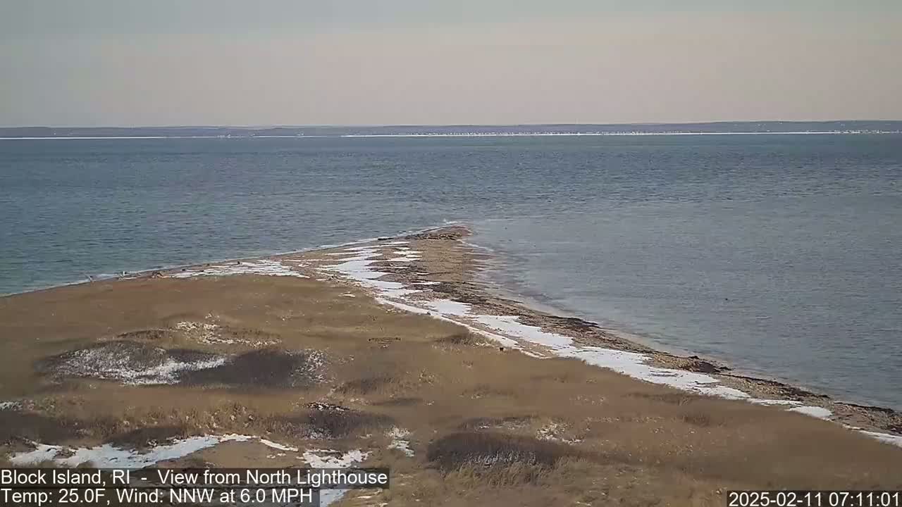 A partially snow-covered, sandy peninsula juts into a calm, gray-blue ocean under a clear, light sky.