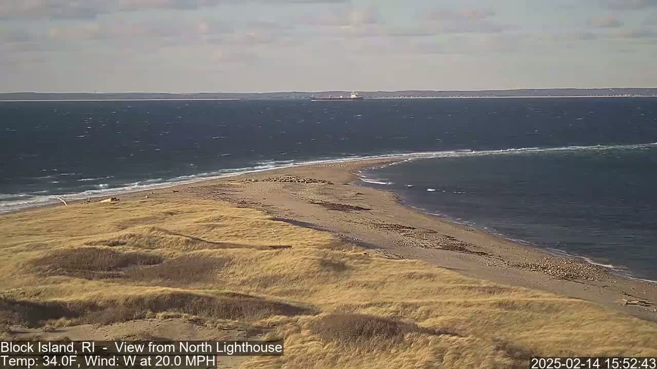 A sandy, grassy point of land juts into a choppy ocean under a partly cloudy sky, with a large ship visible in the distance.