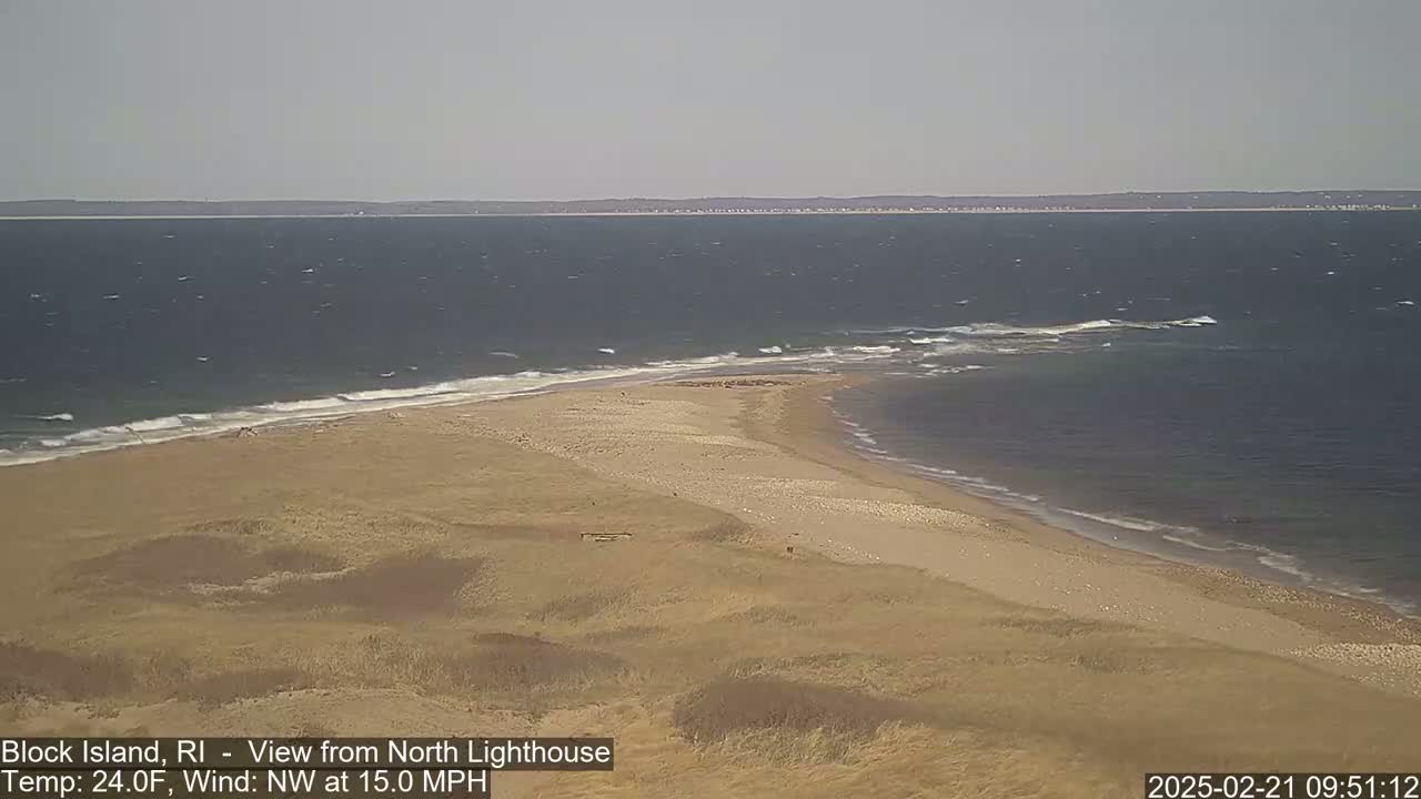 A sandy beach and grassy dunes meet a choppy ocean under a mostly clear sky.