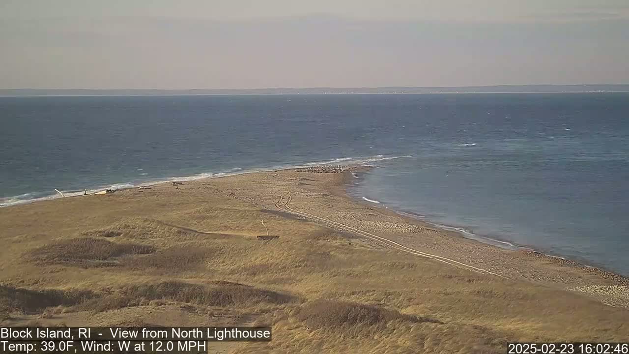 A sandy, dune-covered point of land juts into a blue ocean under a mostly clear sky with a 12 mph westerly wind.