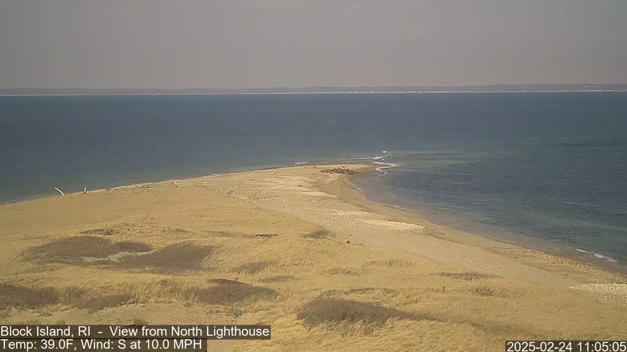 A sandy beach and dune landscape extends into a calm, blue ocean under a hazy sky.