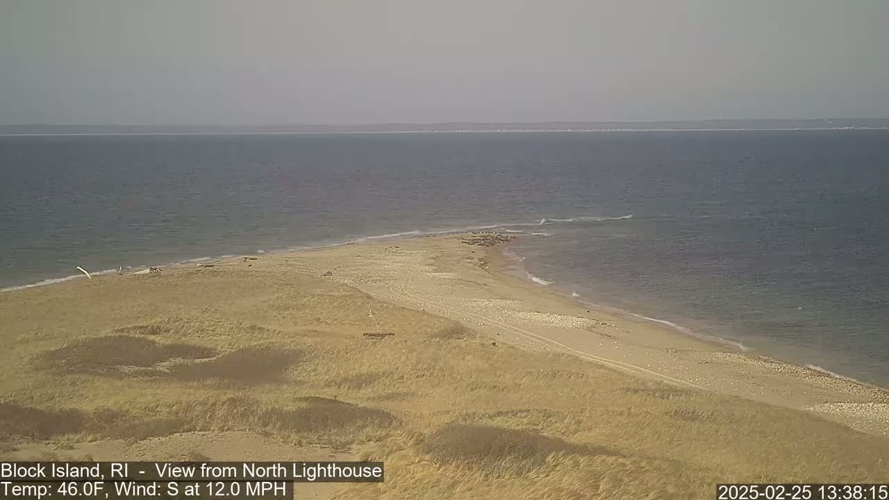 A sandy beach with dunes curves into a calm ocean under a hazy sky.