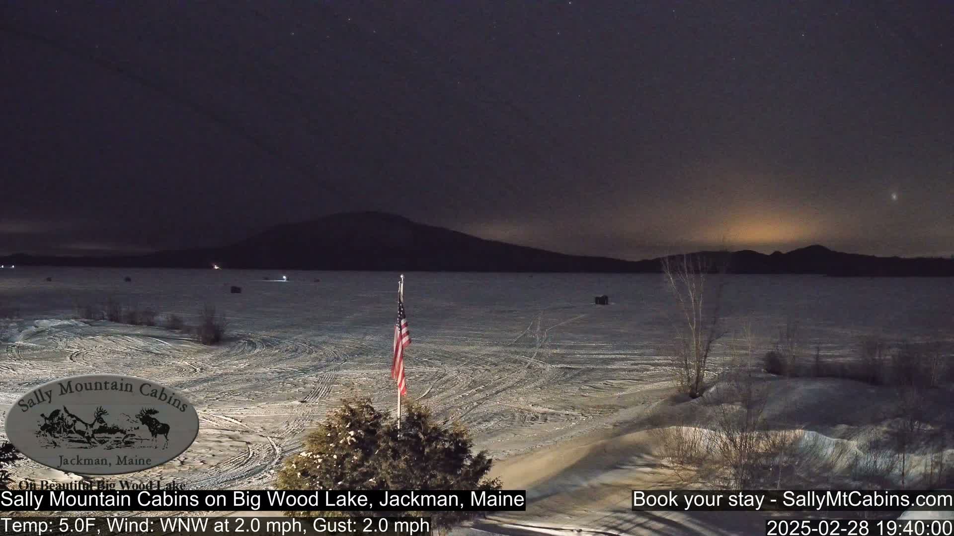 A frozen lake is seen at night under a clear, starry sky, with a distant mountain range and a small American flag in the foreground.
